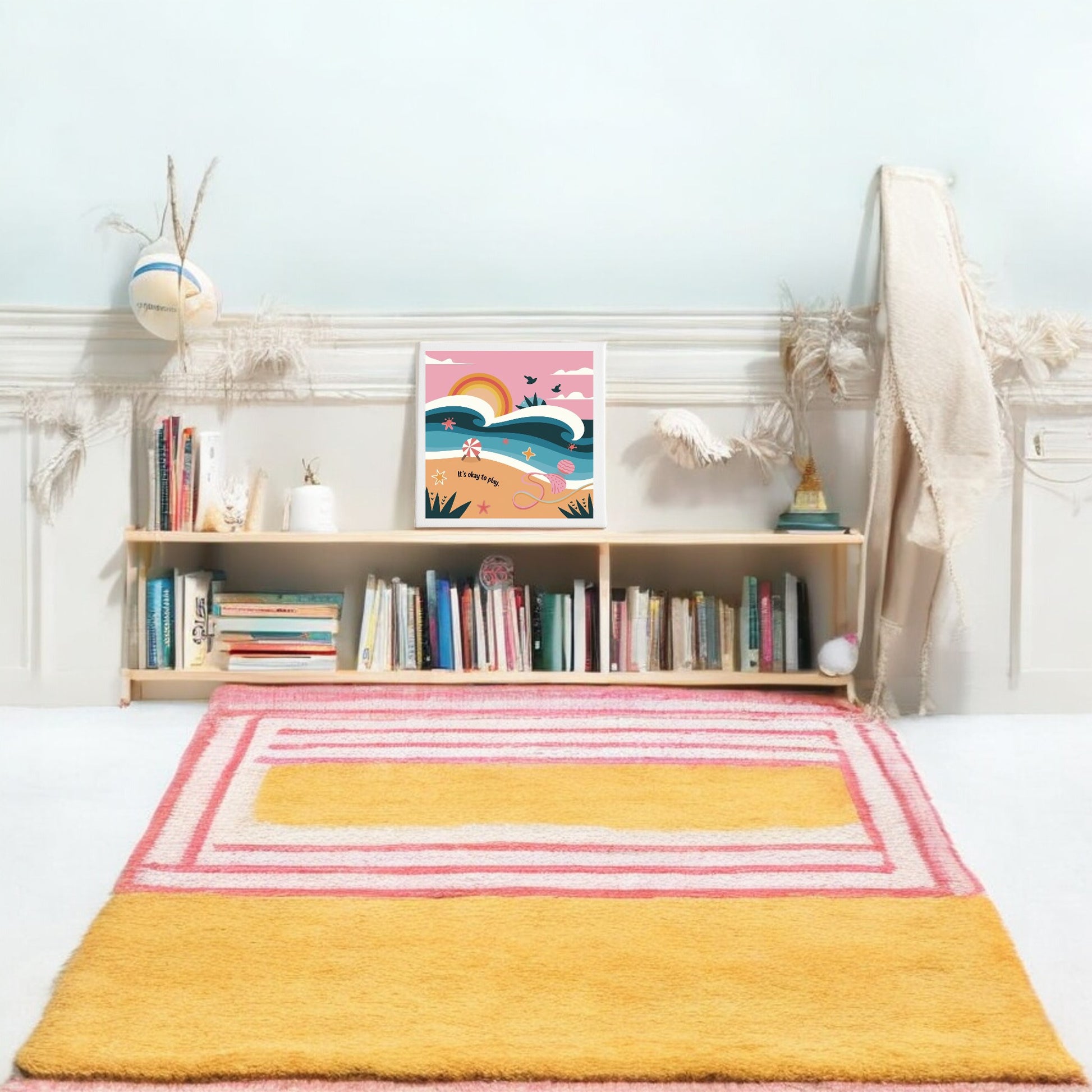 A cozy room with colorful decor features a pale blue wall, a low wooden bookshelf, beach accents, Mirabilia Boutique’s “It’s Okay to Play Canvas – Whimsical Beach Wall Art,” and a yellow and pink geometric rug on the white floor.