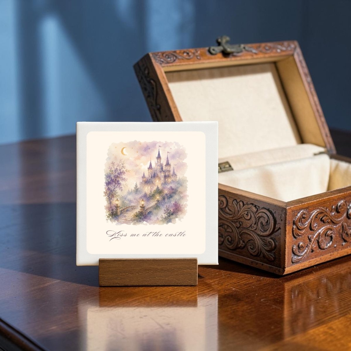 The Kiss Me at the Castle Mini Canvas Sign by Mirabilia Boutique features a pastel castle painting and the phrase "Kiss me at the castle" on a decorative mini canvas with a wooden stand, shown beside an ornate open jewelry box on a polished wooden table—an enchanting fairytale romance accent.