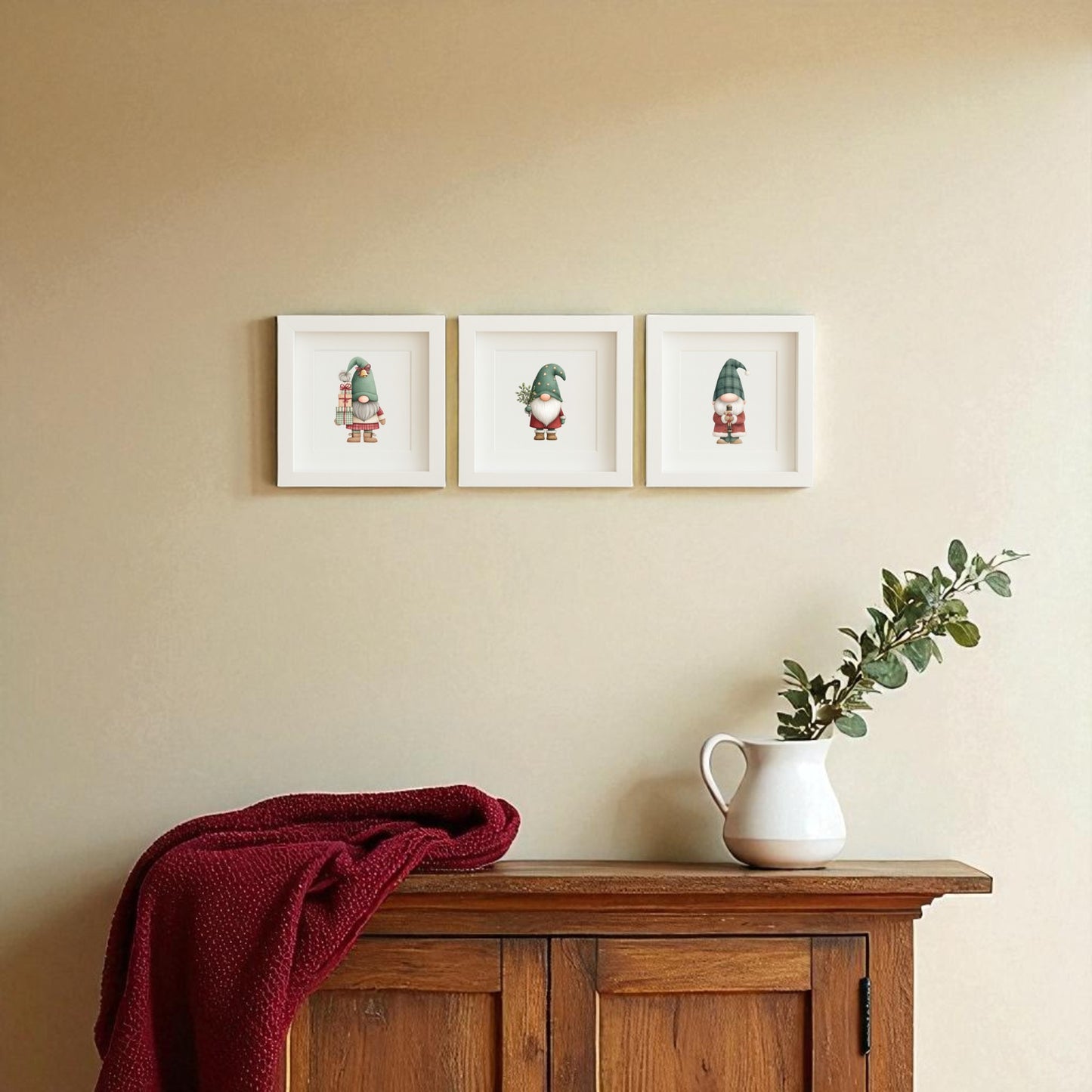 A wooden sideboard with a white pitcher of greenery and a red knit blanket is topped by Mirabilia Boutique’s Scandinavian Gnome Art Trio | Framed Christmas Decor, adding cozy whimsy to the beige wall for festive Scandinavian style.