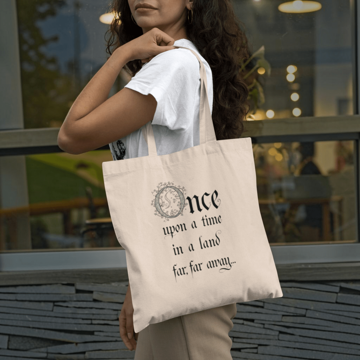 A woman stands holding the "Once Upon a Time Fairytale Tote Bag" by Mirabilia Boutique—a beige literary tote adorned with the phrase "Once upon a time in a land far, far away..." in a whimsical font. She wears a white shirt and is positioned in front of a window and gray stone wall.