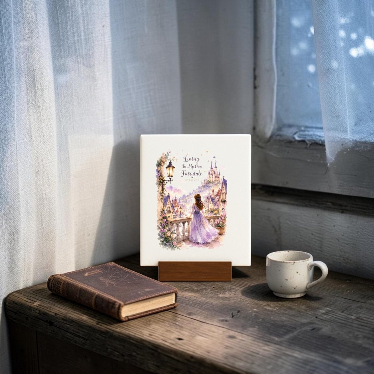 A book and a petite cup sit atop a wooden table next to a whimsical artwork—a Mirabilia Boutique "Living in My Own Fairytale" mini canvas, featuring a princess in a lavender dress standing near a castle with the phrase "Living in My Own Fairytale." Sunlight streams through sheer white curtains, enhancing the cozy cottagecore atmosphere.