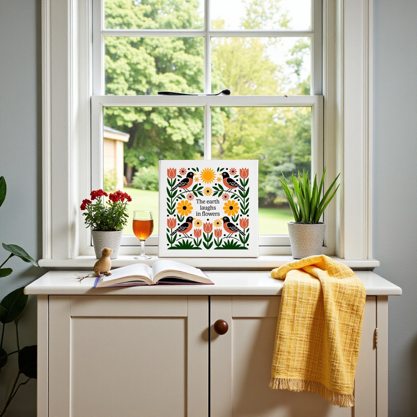 A cozy windowsill with potted plants, tea, an open book, a yellow cloth, and the Mirabilia Boutique "The Earth Laughs in Flowers" Canvas Wall Art featuring floral designs, overlooking a green garden.