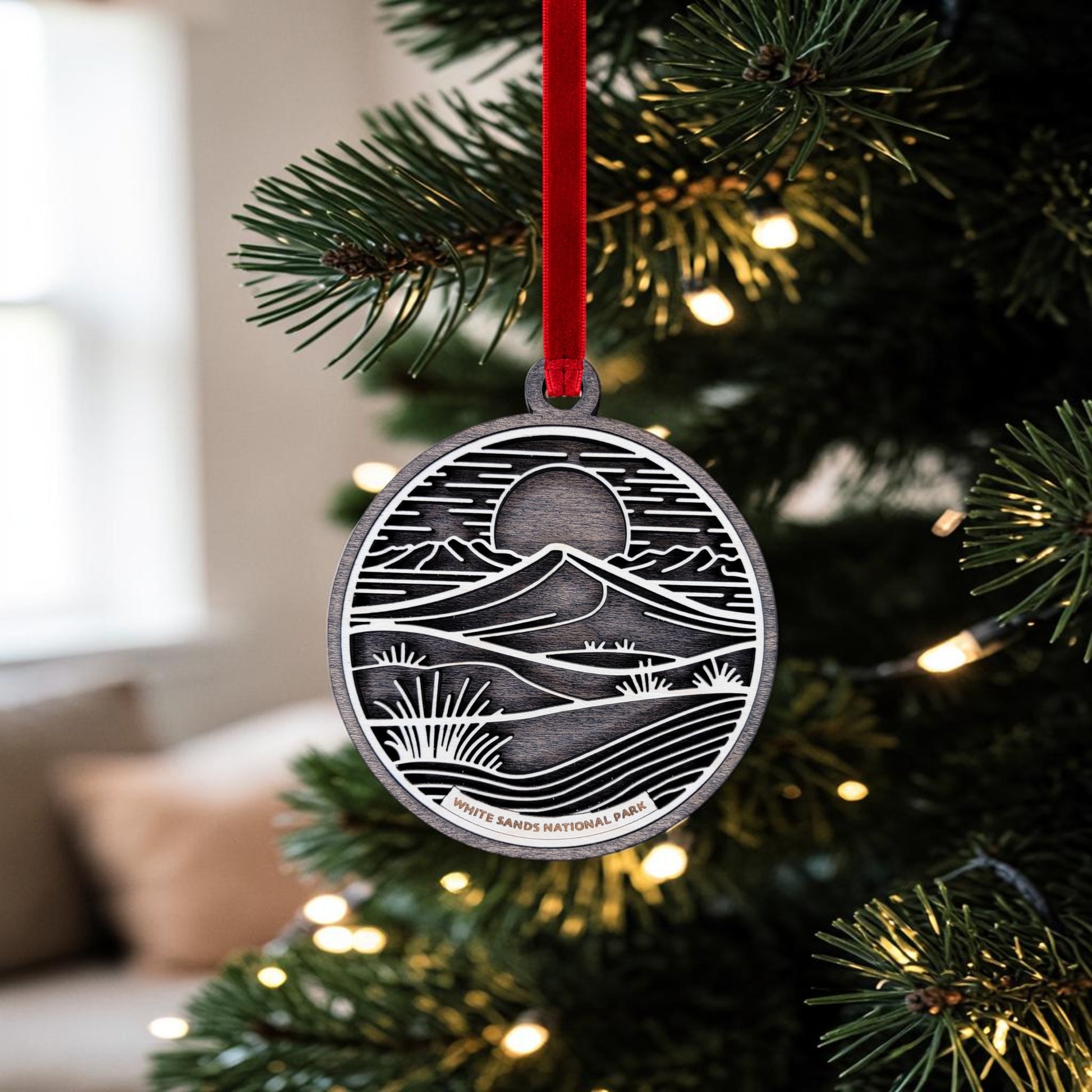 The White Sands National Park Line Art Christmas Ornament by Mirabilia Boutique, handcrafted in an oval wooden shape, hangs from a red ribbon on a Christmas tree adorned with warm lights. The ornament displays an engraved landscape of gypsum dunes, mountains, sun, and desert plants with “White Sands National Park” etched at the bottom.