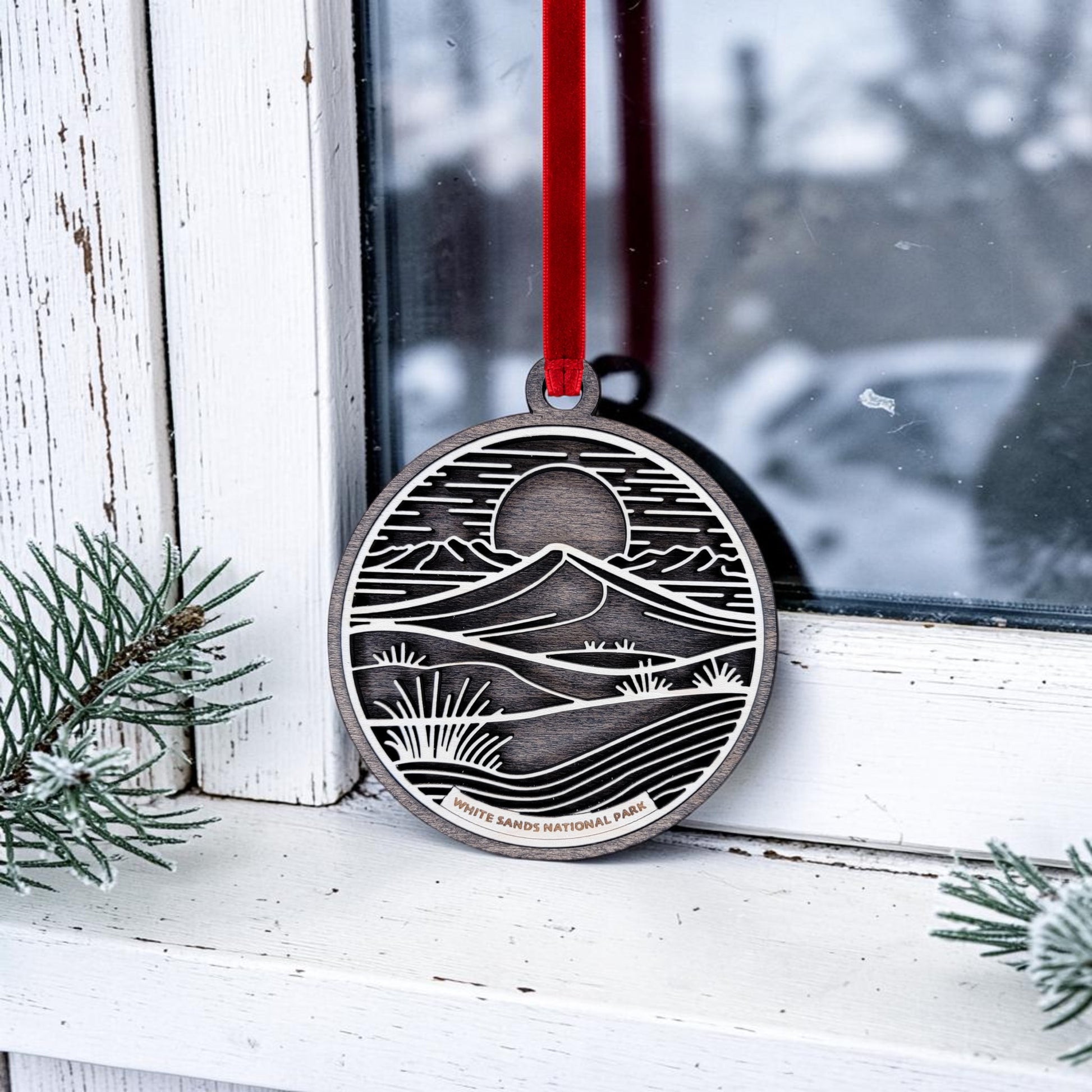 The Mirabilia Boutique White Sands National Park Line Art Christmas Ornament, featuring a mountain and sun design inspired by the gypsum dunes of White Sands National Park, hangs by a red ribbon in a window. “White Sands National Park” is engraved at the bottom, while snowy pine branches and frost frame the scene.