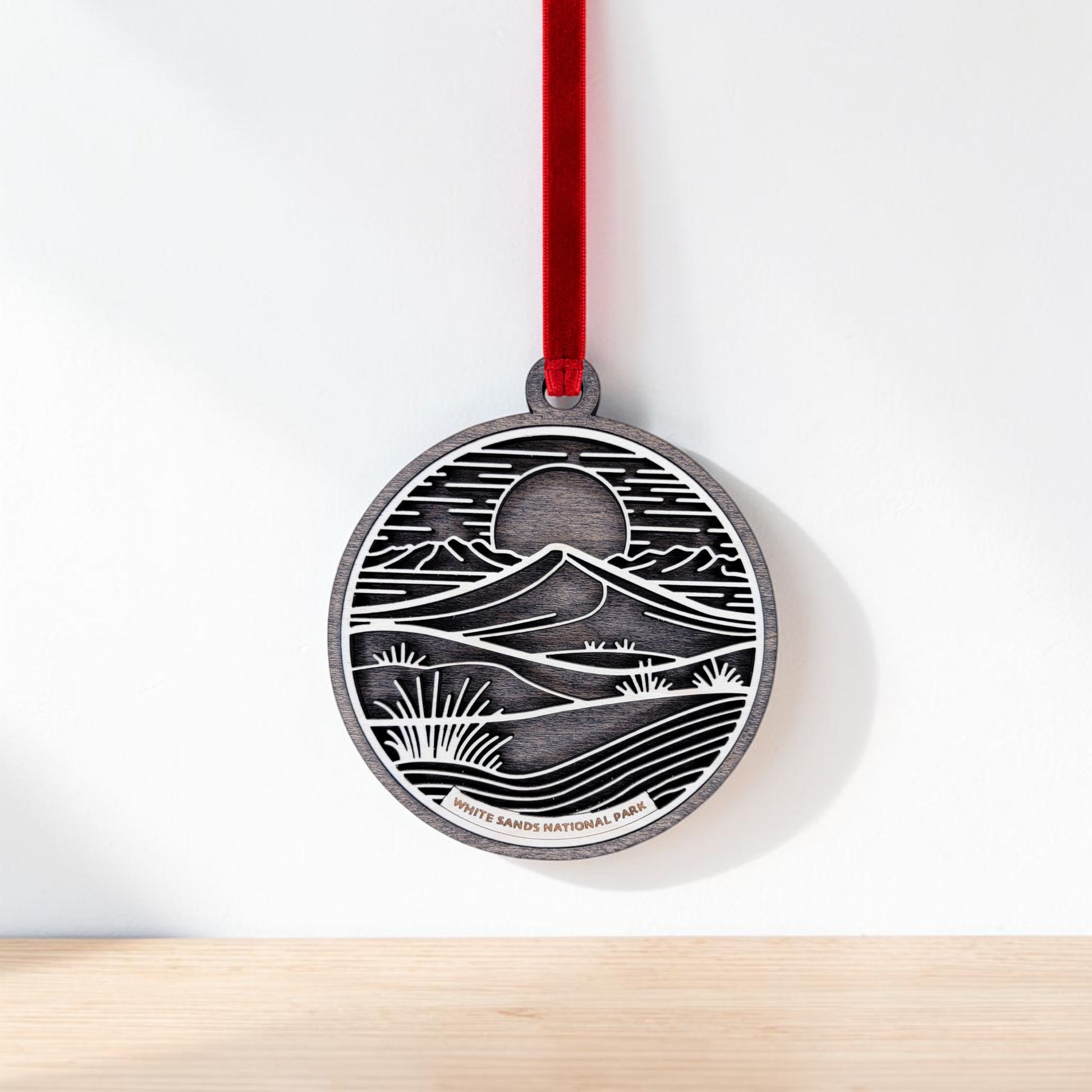 The Mirabilia Boutique White Sands National Park Line Art Christmas Ornament is a handcrafted wooden decoration featuring an engraved desert landscape with gypsum dunes, mountains, plants, a sun or moon, and the words "White Sands National Park." It hangs from a red ribbon and is displayed against a white background.