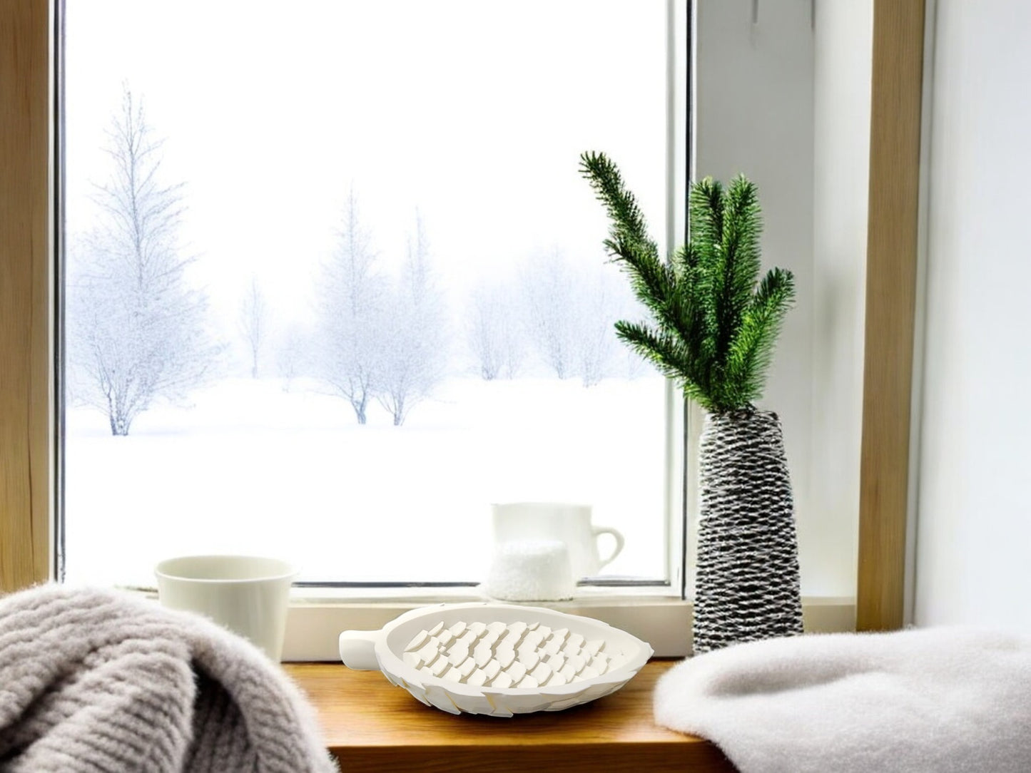 A cozy windowsill features the Mirabilia Boutique Winter White Pinecone Tray for Woodland Winter Decor, paired with a white mug, pitcher, knotted vase with an evergreen branch, and blanket. Snow-covered trees outside complete the winter scene.