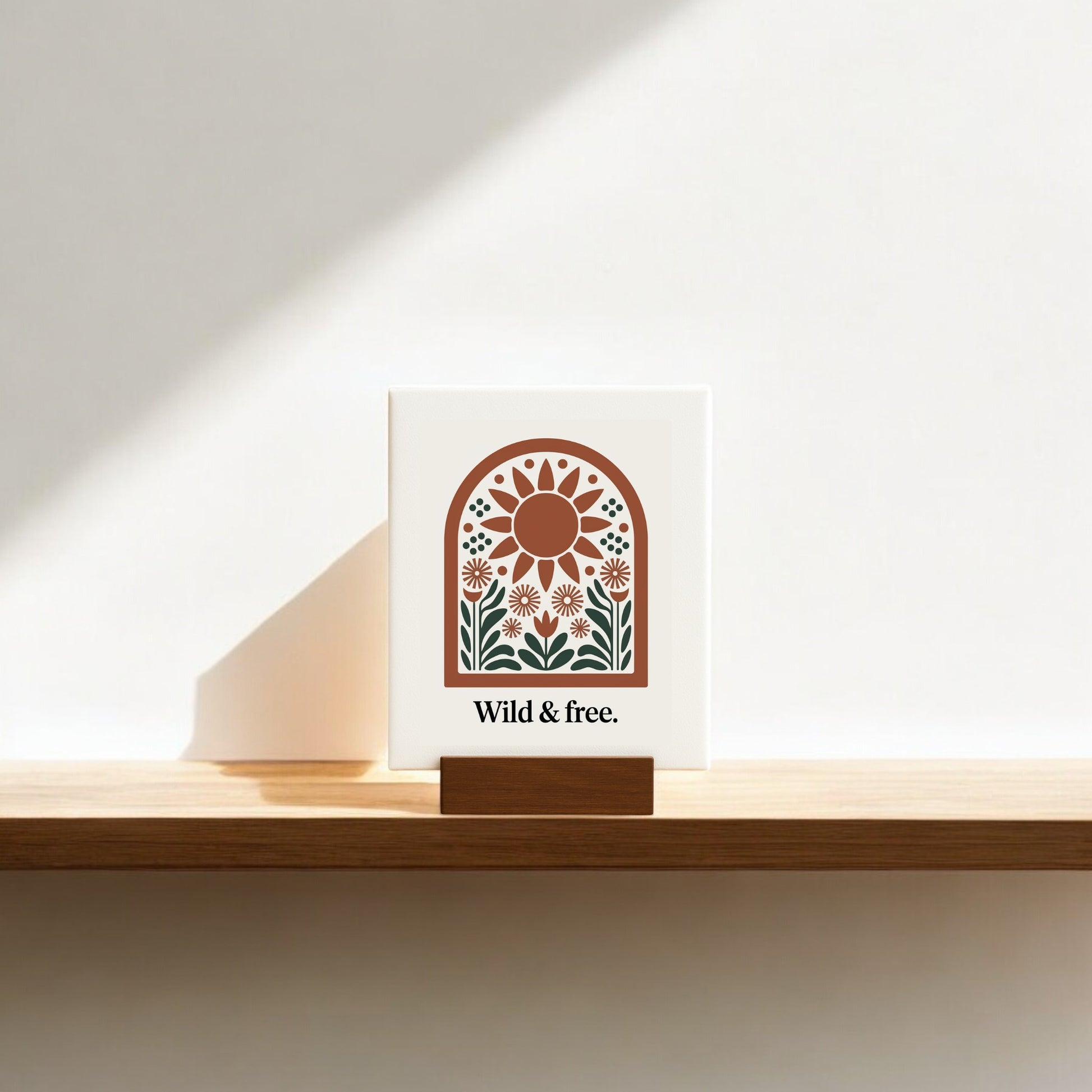 The Wild & Free Mini Sign – Sunflower Boho Desk Art by Mirabilia Boutique sits on a wooden stand, its earthy sun and floral design casting a soft shadow for an inviting boho vibe. Perfect shelf or desk decor.