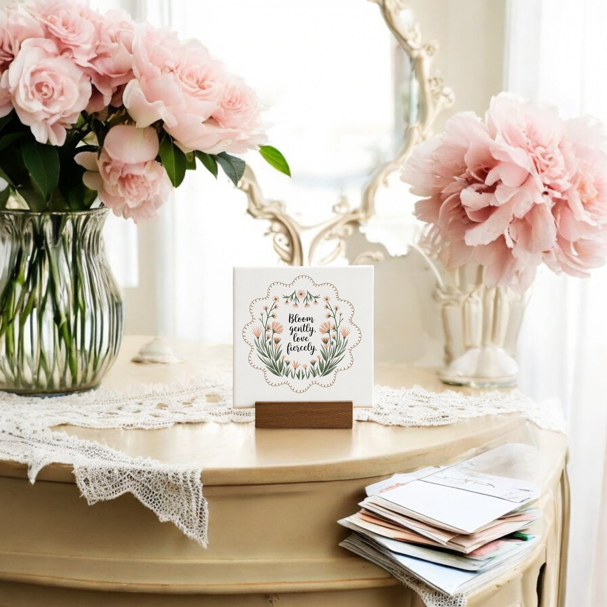 The Bloom Gently Love Fiercely Mini Canvas Sign by Mirabilia Boutique sits on a vintage vanity with lace, pink peonies, a mirror, and stacked envelopes—a charming encouragement gift for wildflower decor lovers.