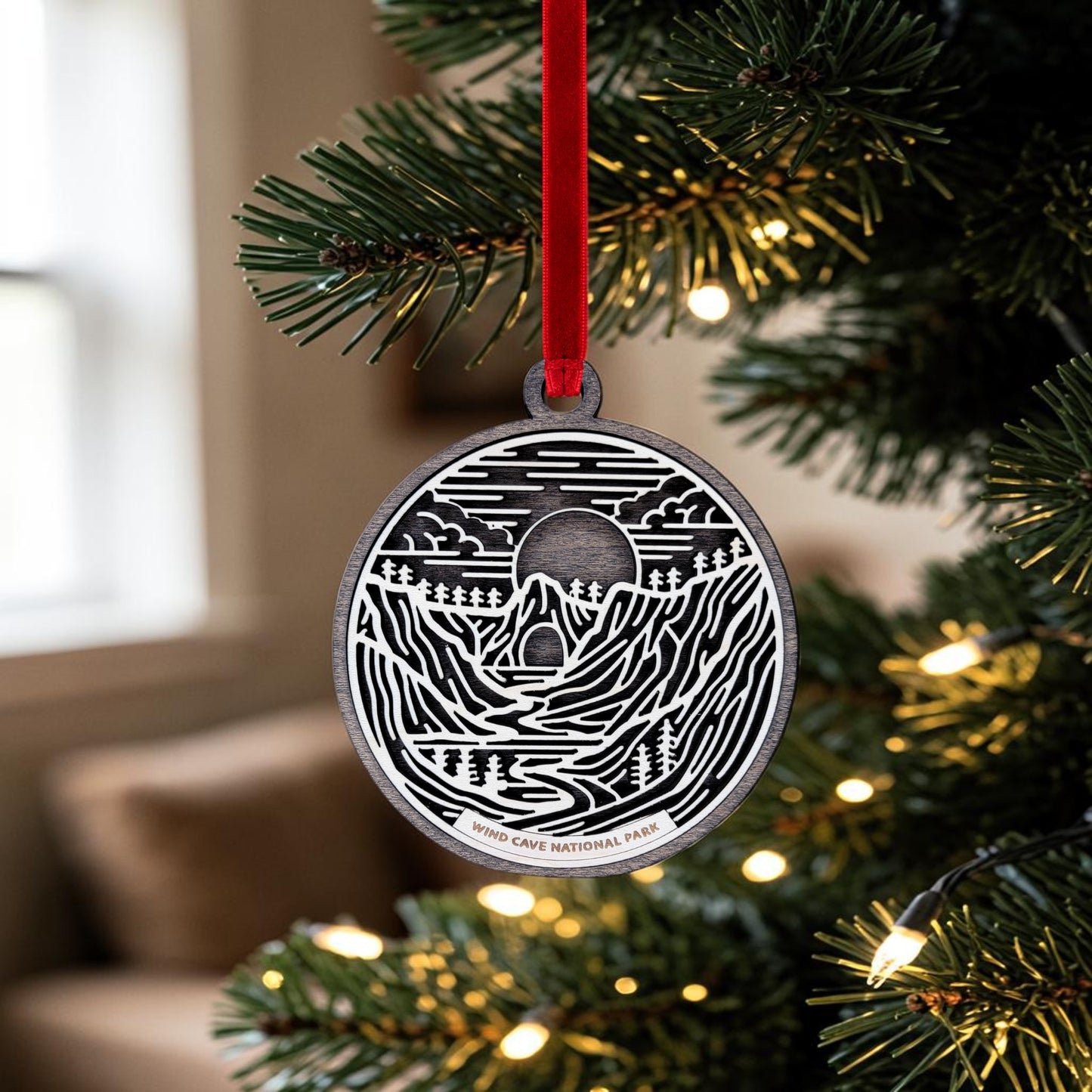 The Mirabilia Boutique Wind Cave National Park Line Art Christmas Ornament, featuring a round metal design with landscape artwork and "Wind Cave National Park" text, hangs from a red ribbon on a Christmas tree adorned with lights, celebrating the natural beauty of the Black Hills.