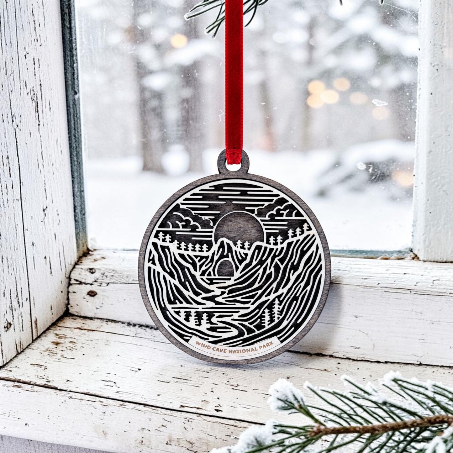 A Wind Cave National Park Line Art Christmas Ornament by Mirabilia Boutique, showcasing a detailed nature scene and the park’s name, hangs by a red ribbon on a snowy windowsill, evoking the tranquil beauty of the Black Hills with pine branches and snow framed outside.