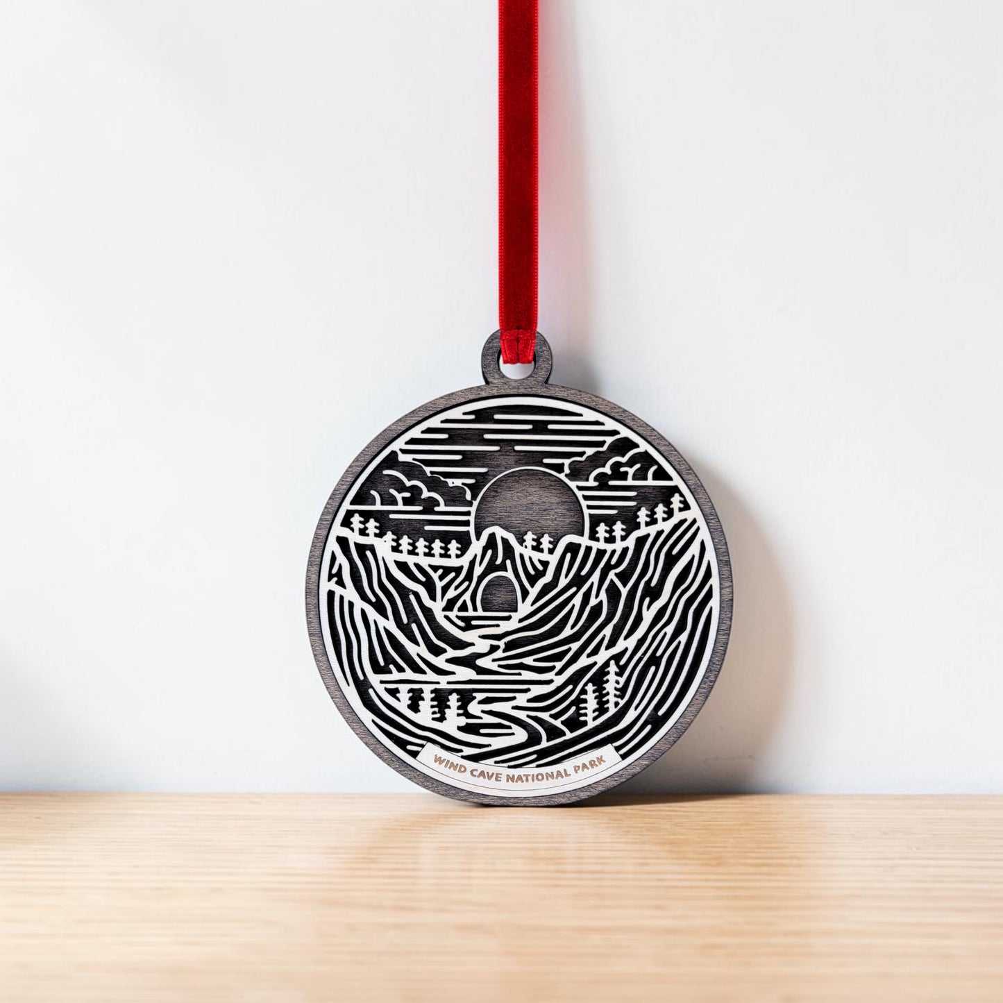 The Wind Cave National Park Line Art Christmas Ornament by Mirabilia Boutique is a handcrafted wooden ornament featuring a round design with mountain, cave, river, and tree motifs inspired by the scenic Black Hills. Hanging from a red ribbon, "Wind Cave National Park" appears beneath the design. The ornament is pictured against a white background and wooden surface.