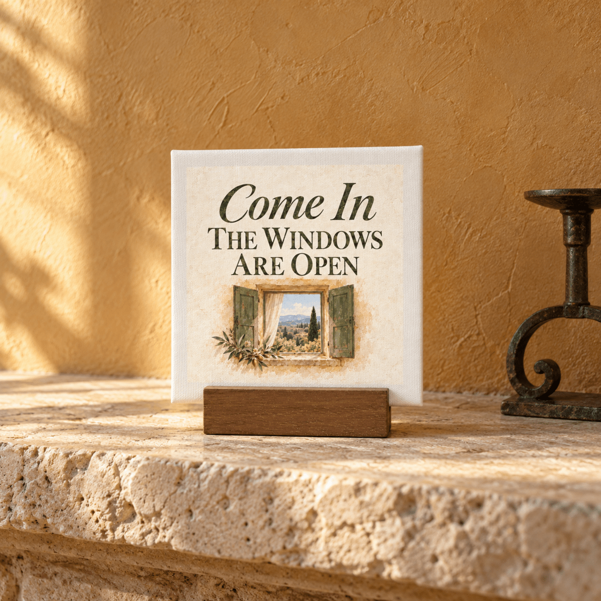 A Come In Mini Canvas Sign | Ochre & Cypress on a wooden stand reads, “Come In The Windows Are Open,” featuring a painted illustration of an open window overlooking a countryside landscape.