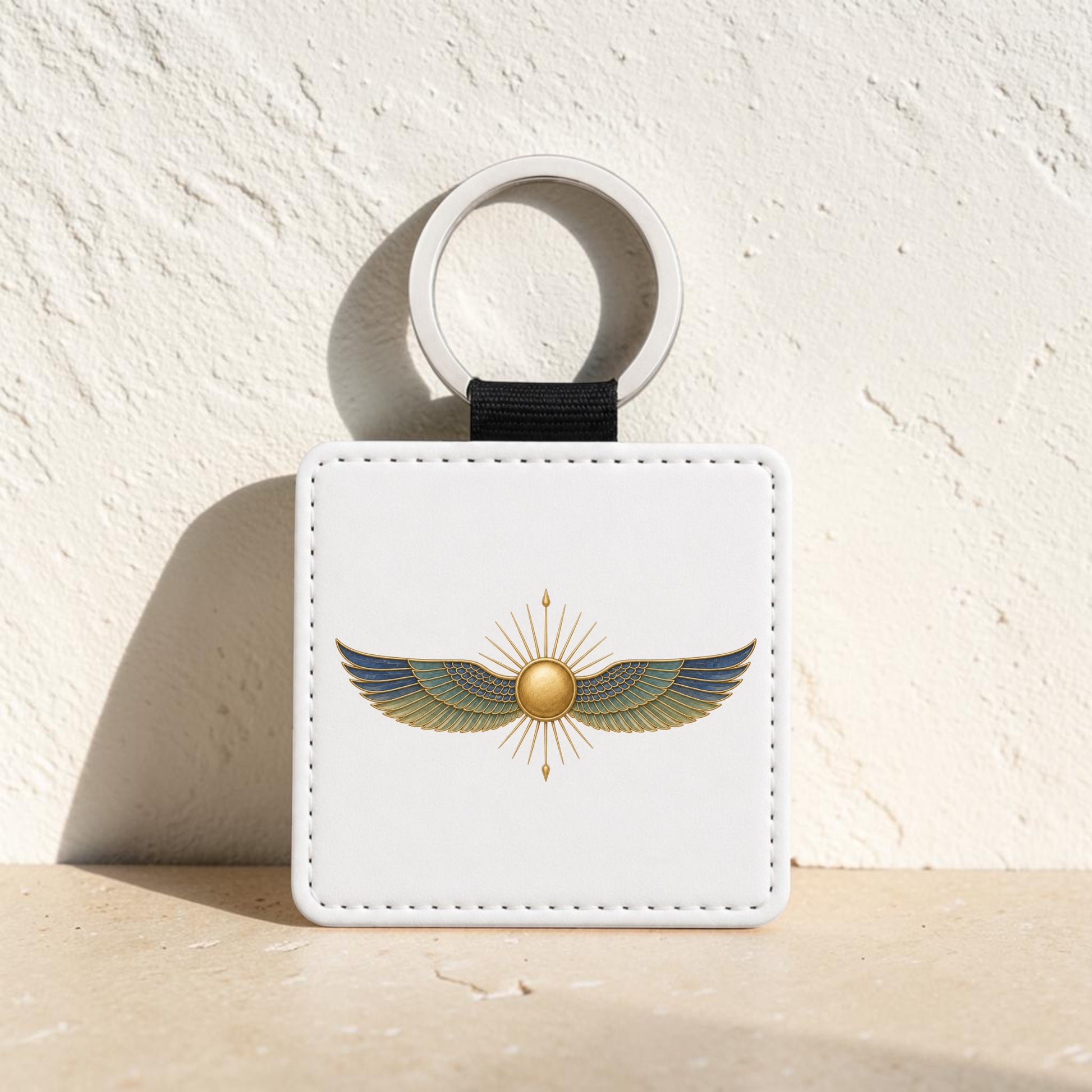 Introducing the Winged Sun Keychain | Egyptian Inspired Keepsake from Mirabilia Boutique’s Lapis & Lotus Collection—a white square decorative keychain featuring a gold and green Egyptian-style winged sun design, displayed against a textured beige surface with sunlight creating a dramatic shadow behind it.