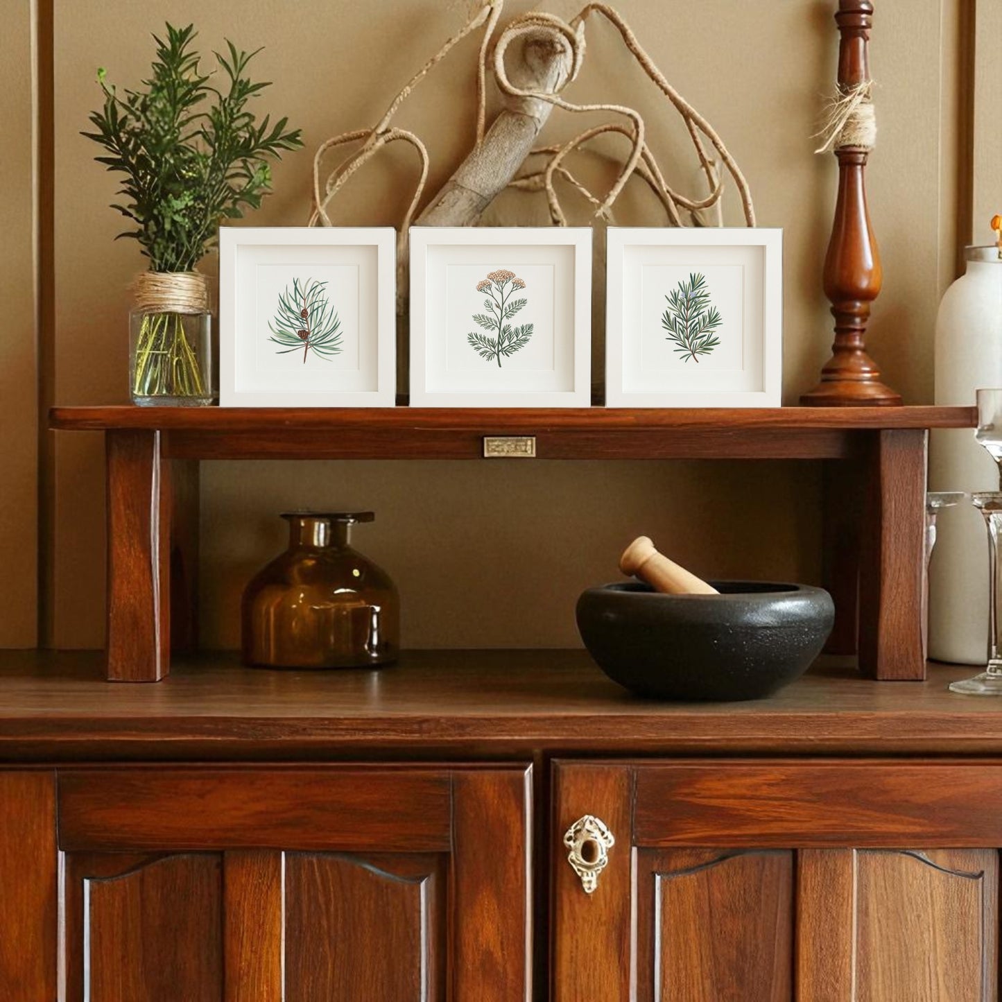 A wooden sideboard features the Winter Botanical Trio | Pine Yarrow Rosemary Prints by Mirabilia Boutique, displayed in frames with a vase of greenery, brown jar, mortar and pestle, candlestick, and rope decor against a tan wall.