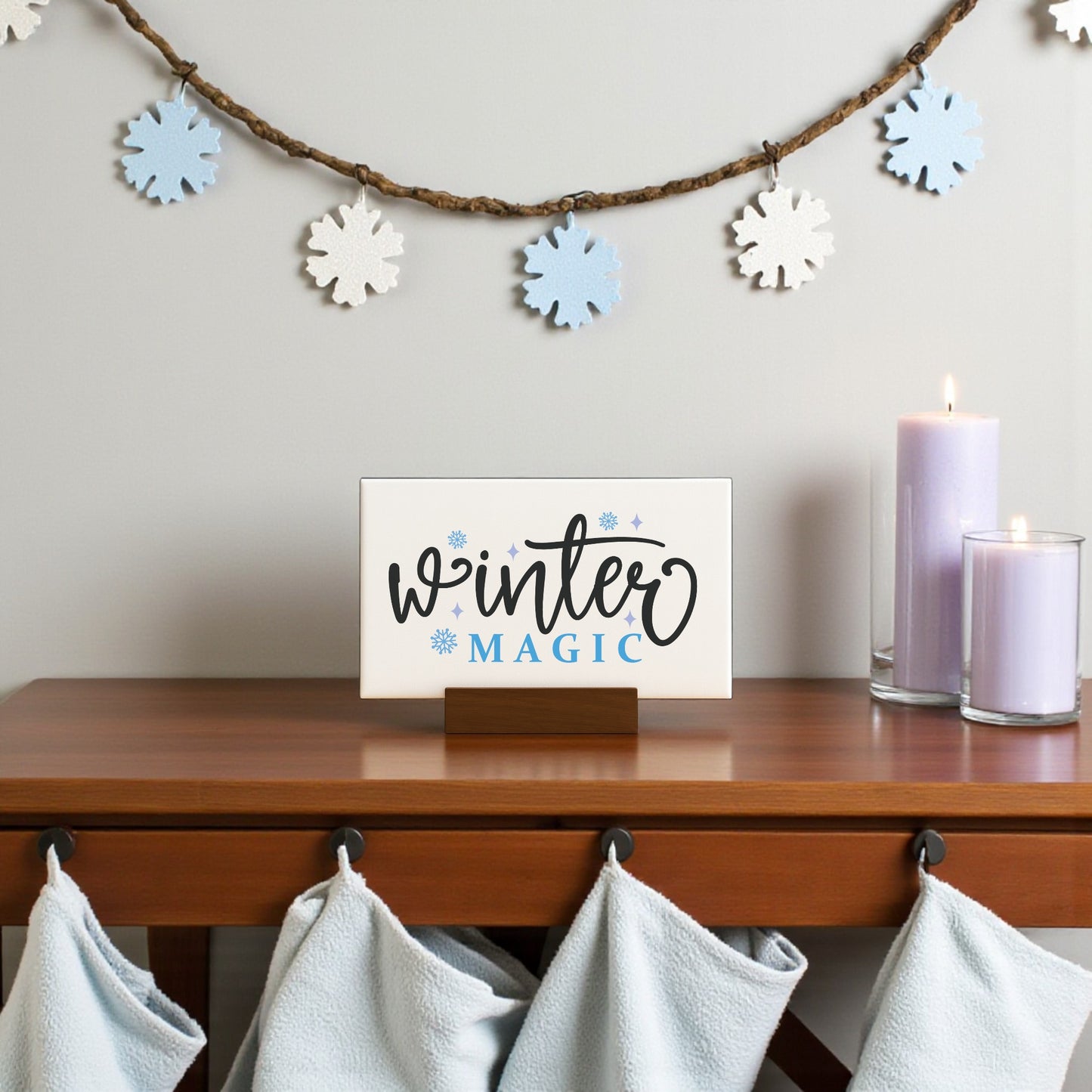 A Mirabilia Boutique Winter Magic Sign (6x4 Minimalist Winter Decor) sits on a wooden table beside two lit lavender candles. Blue and white snowflake garland hangs above, with light blue towels on hooks below.