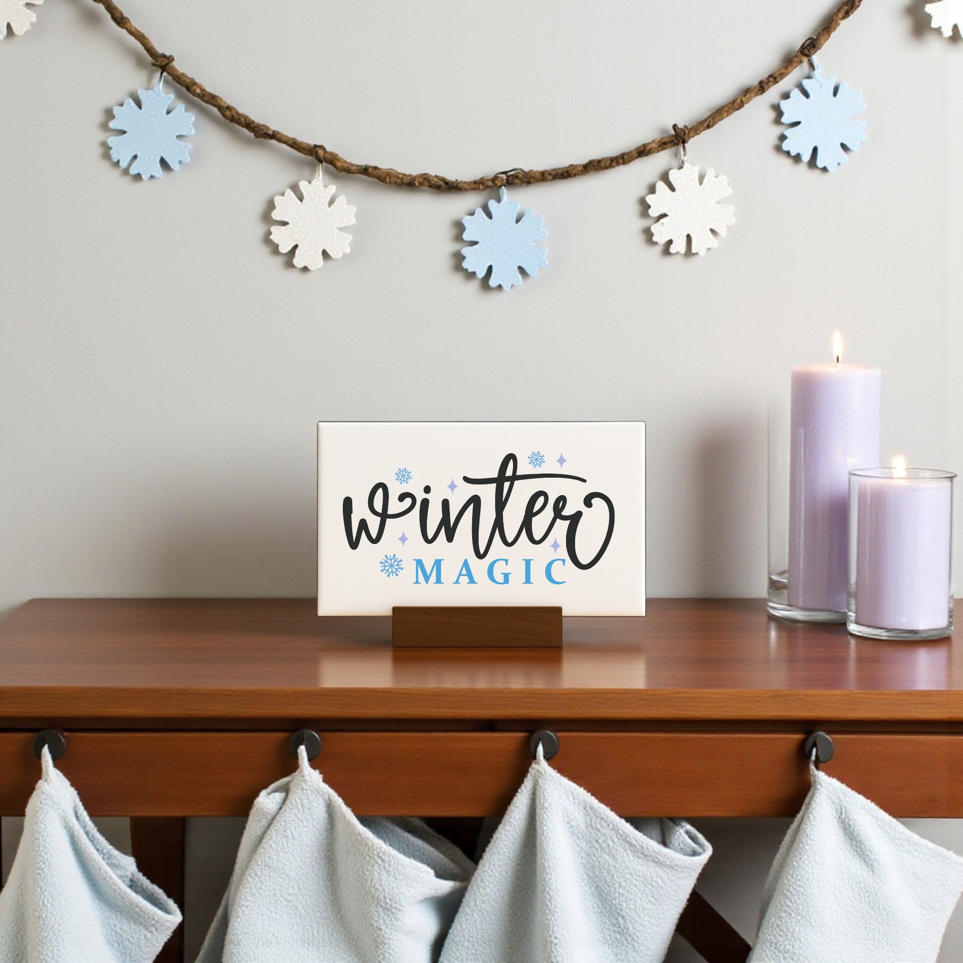 A Mirabilia Boutique Winter Magic Sign (6x4 Minimalist Winter Decor) sits on a wooden table beside two lit lavender candles. Blue and white snowflake garland hangs above, with light blue towels on hooks below.