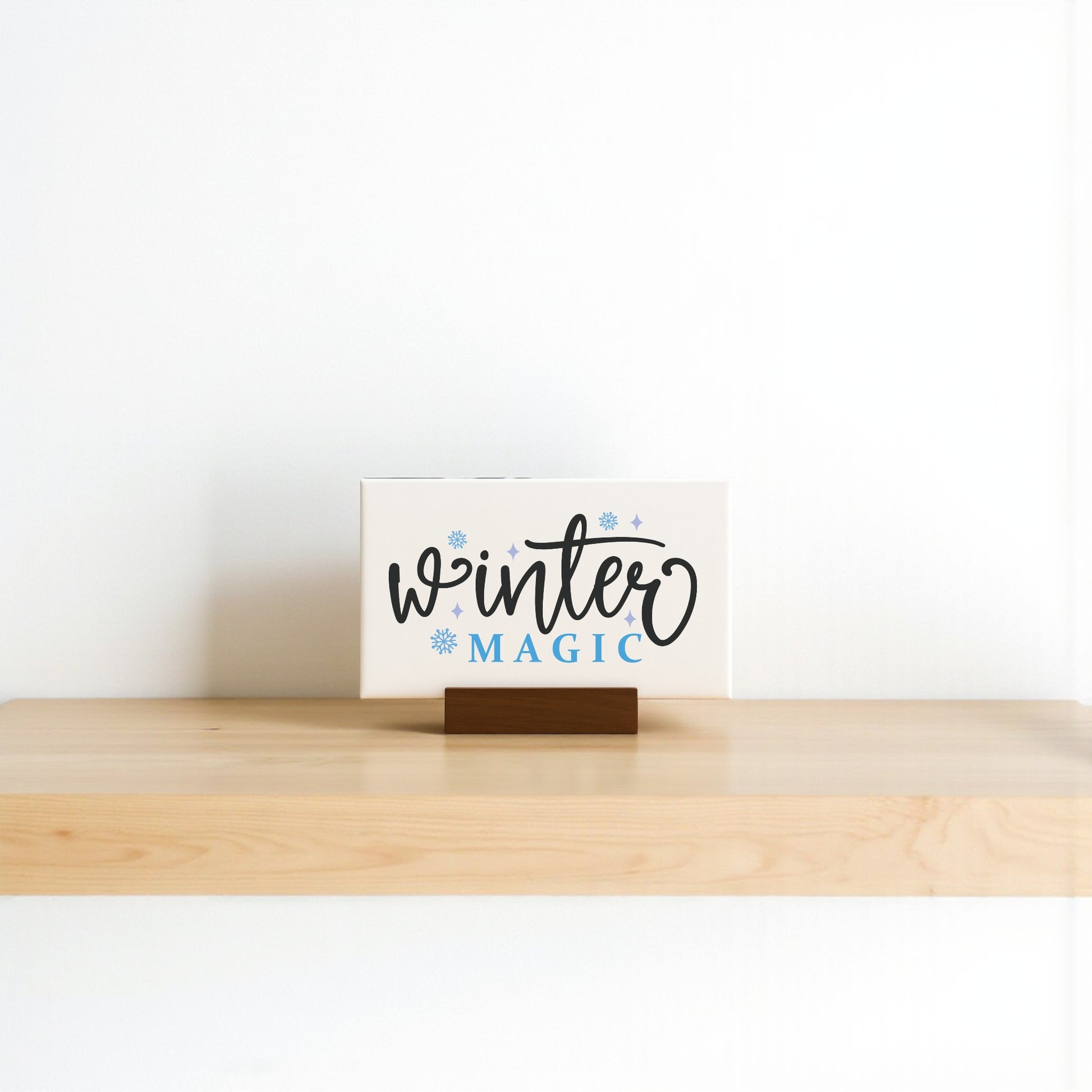 A 6x4 Winter Magic Sign from Mirabilia Boutique, featuring decorative lettering and blue snowflakes, sits on a wooden shelf, adding the perfect minimalist seasonal touch to your home against a plain white background.