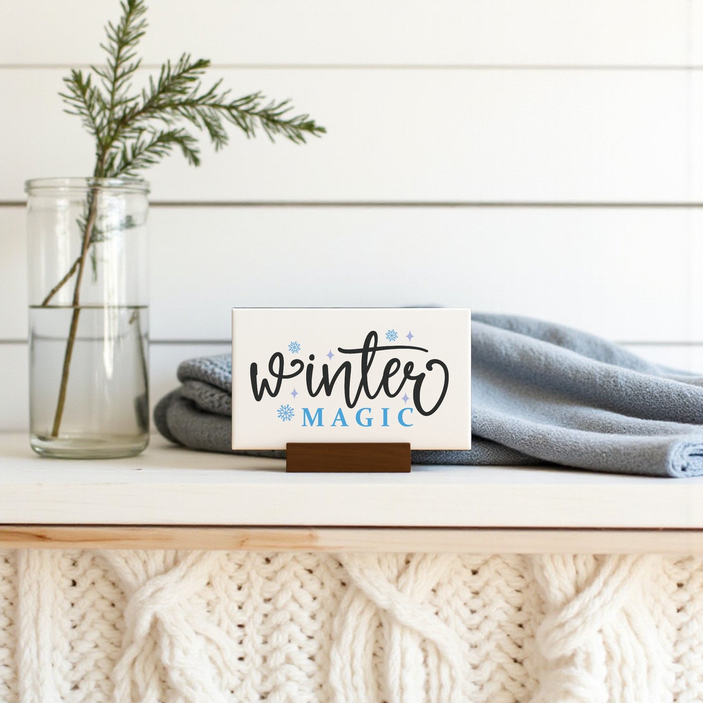 The Mirabilia Boutique Winter Magic Sign | 6x4 Minimalist Winter Decor sits on a white mantel with a knit pattern, beside a glass jar of evergreens and a folded light blue blanket—perfect for cozy seasonal home accents.