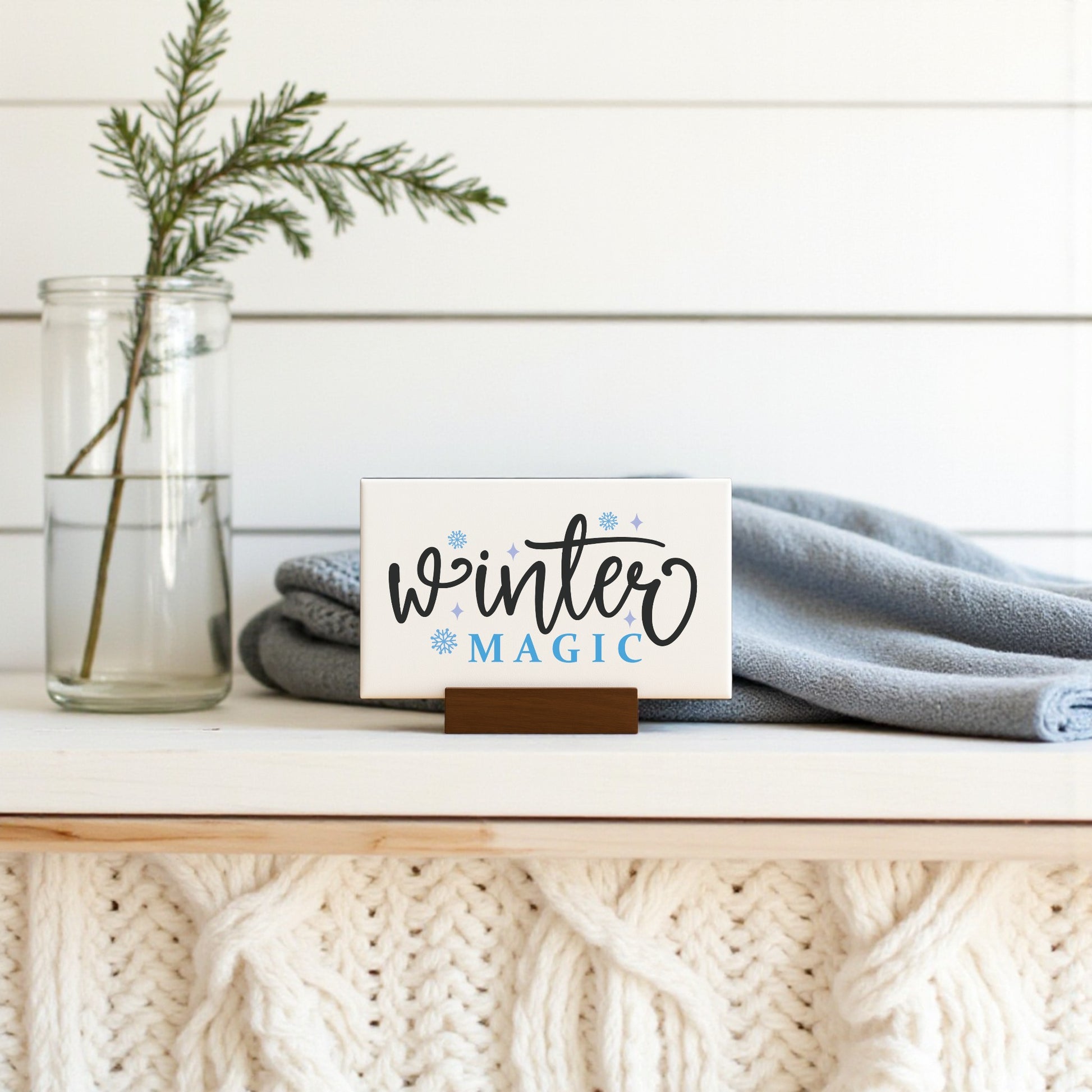 The Mirabilia Boutique Winter Magic Sign | 6x4 Minimalist Winter Decor sits on a white mantel with a knit pattern, beside a glass jar of evergreens and a folded light blue blanket—perfect for cozy seasonal home accents.