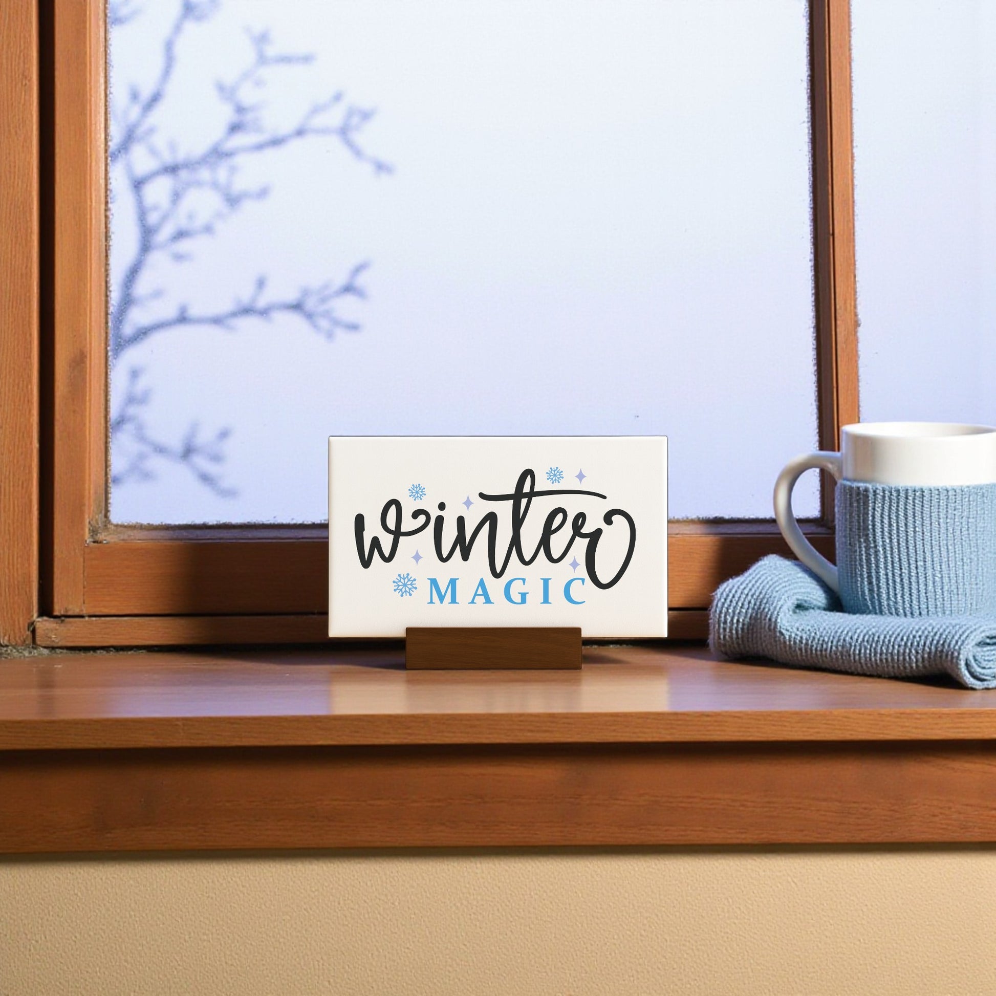 The Winter Magic Sign (6x4) by Mirabilia Boutique sits on a wooden windowsill beside a blue knit cloth and white mug, creating cozy minimalist winter decor against a frosty window and bare tree branches.