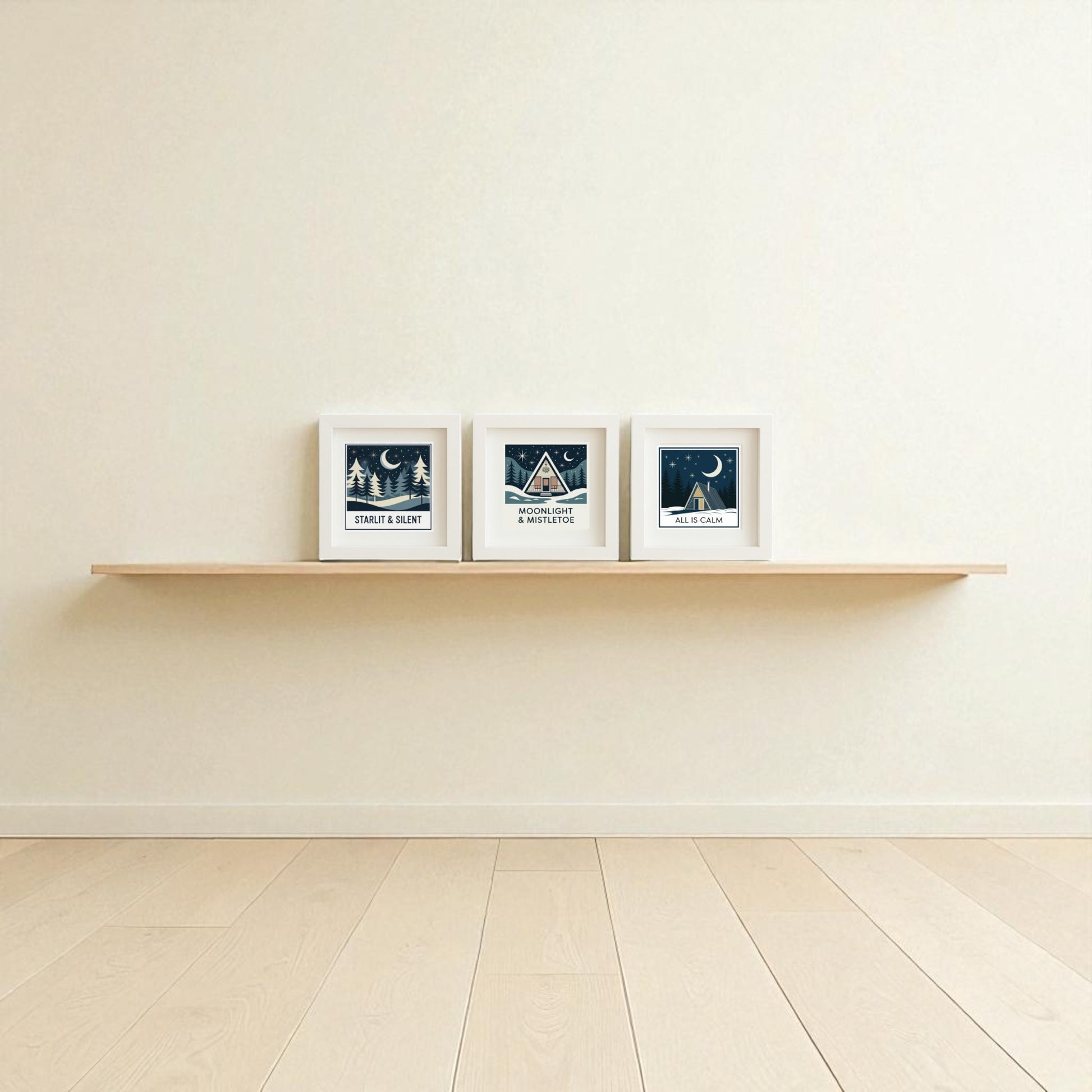 A minimalist room with light wood flooring and cream walls features the Winter Mid-Century Art Trio by Mirabilia Boutique—a floating wooden shelf displays three framed, dark blue and white nature and night sky prints, perfect for cozy holiday wall decor.