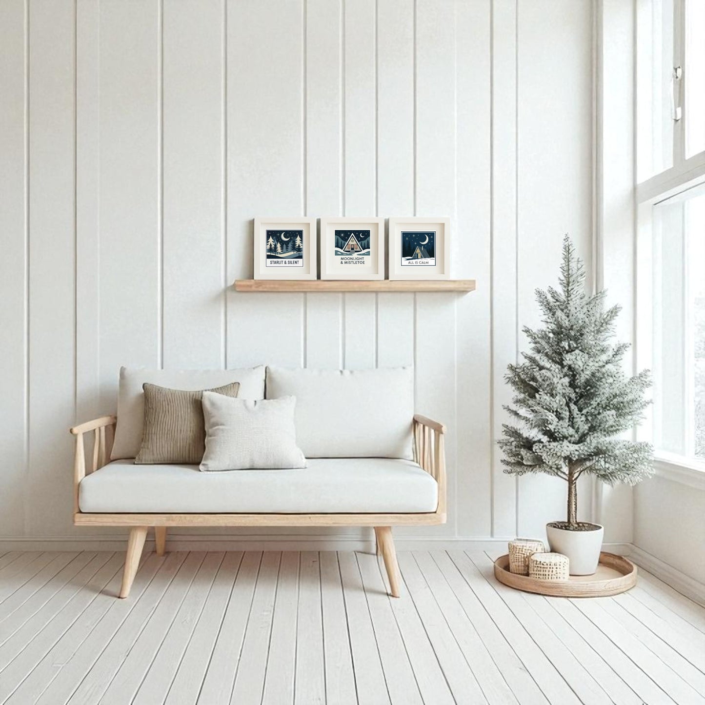 A minimalist living room features a light wood sofa, neutral cushions, and Mirabilia Boutique’s Winter Mid-Century Art Trio wall decor above. A small frosted pine tree adds cozy seasonal charm by the large, sunlit windows.