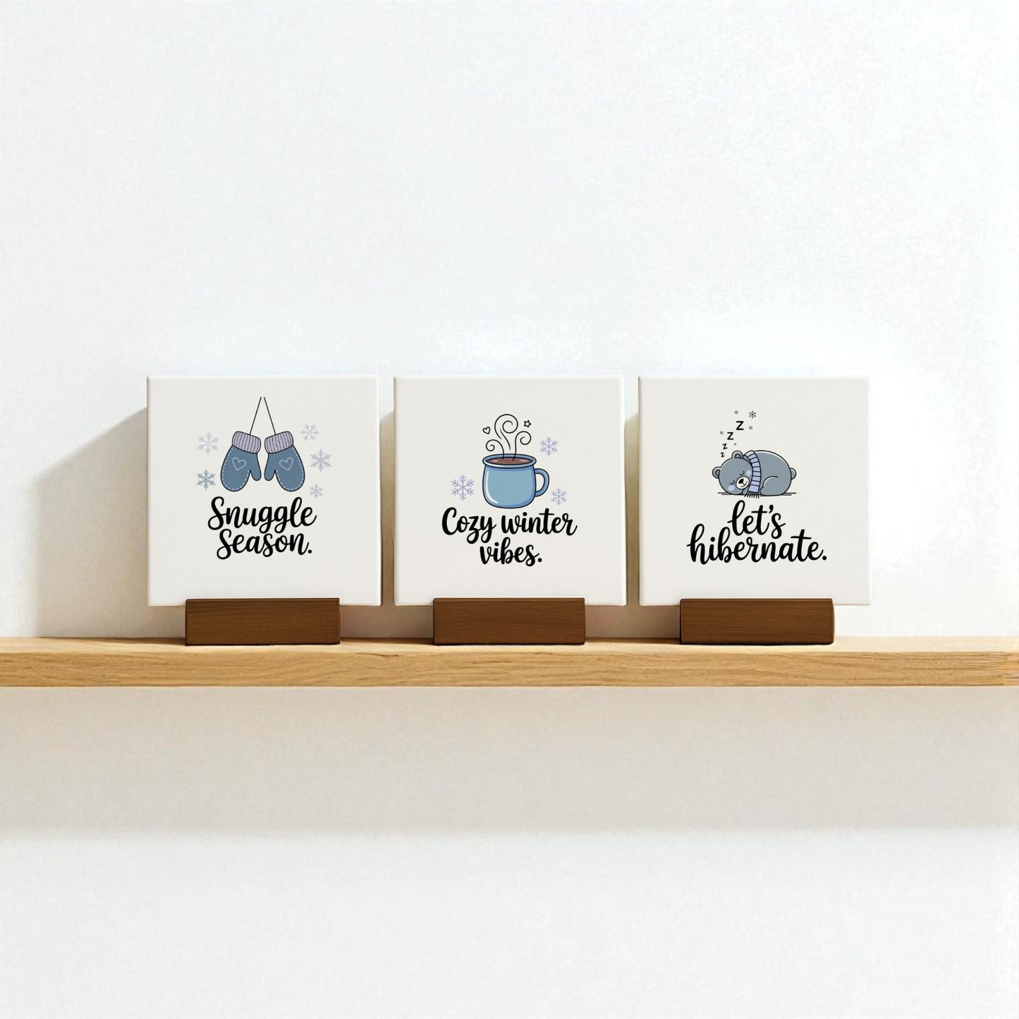 The Winter Canvas Sign Trio by Mirabilia Boutique features three small square signs—mittens with "Snuggle season," a mug with "Cozy winter vibes," and a sleeping bear with "let’s hibernate." Perfect cozy winter decor or charming gift!.