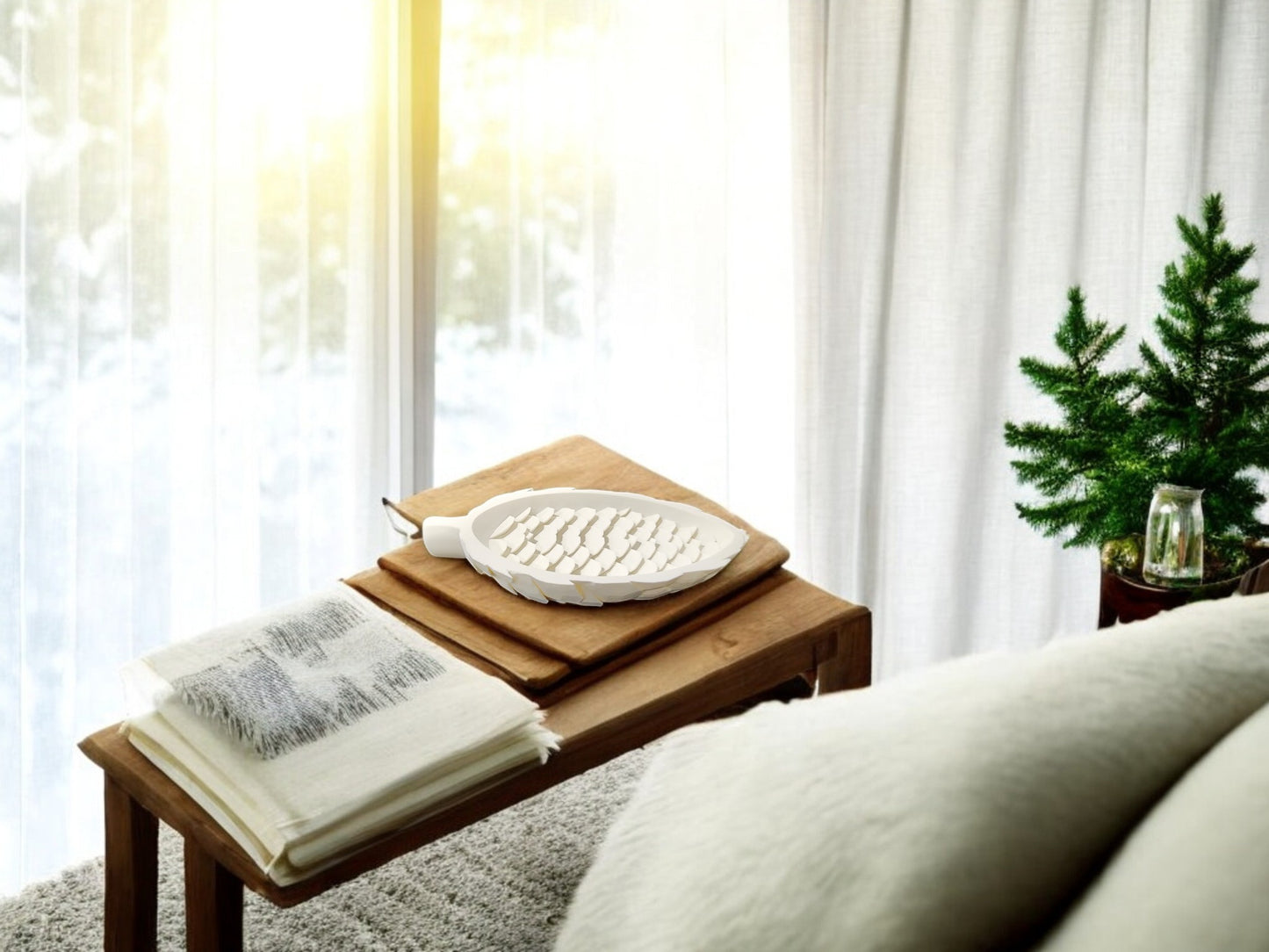 A Mirabilia Boutique Winter White Pinecone Tray sits on a wooden side table with a folded blanket, while a small potted pine adds woodland winter decor. Sunlight through sheer curtains completes the cozy scene.