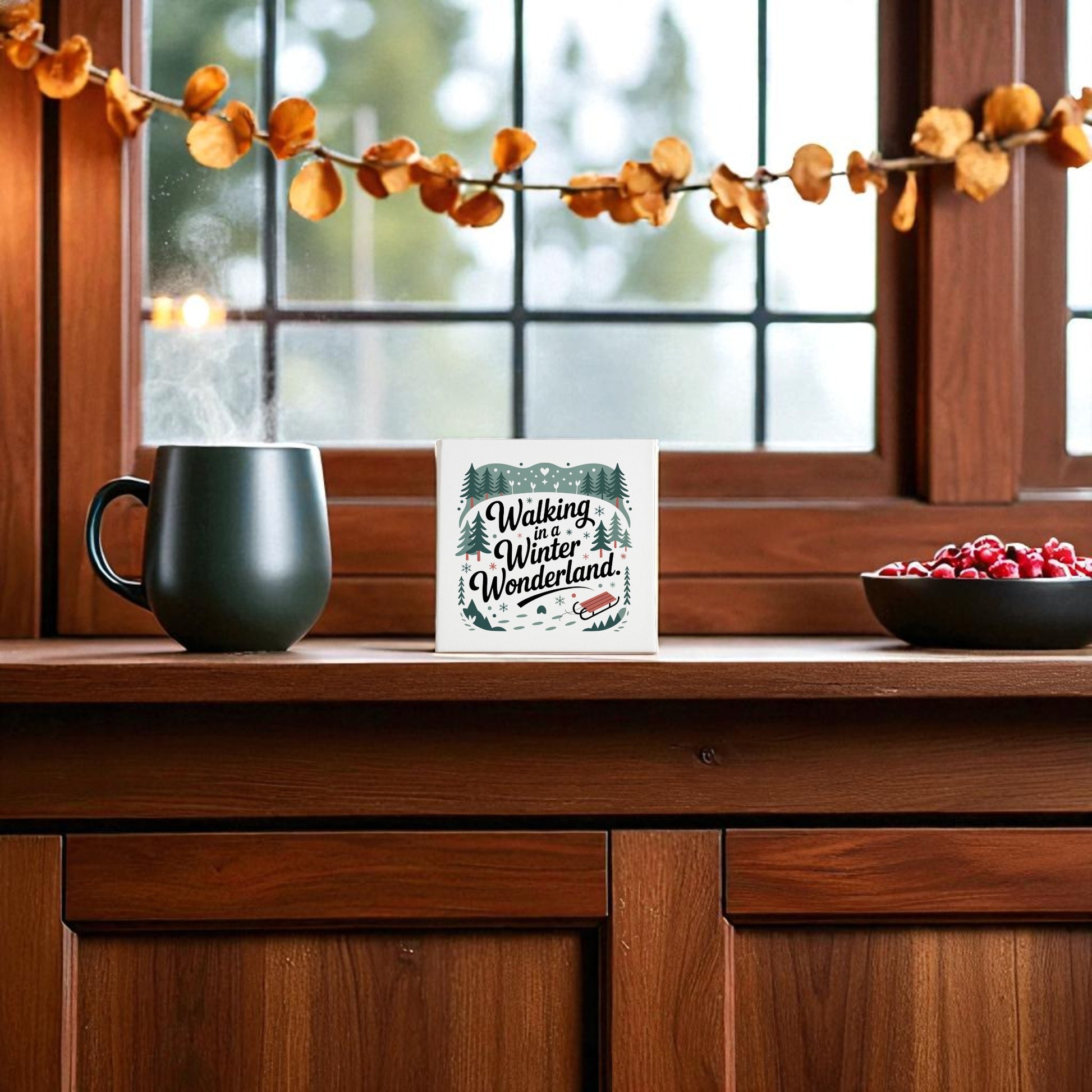 A steaming mug, a bowl of cranberries, and the Mirabilia Boutique "Winter Wonderland Canvas | 8x8 Holiday Decor" sit on a wooden countertop by a window decorated with a dried leaf garland.
