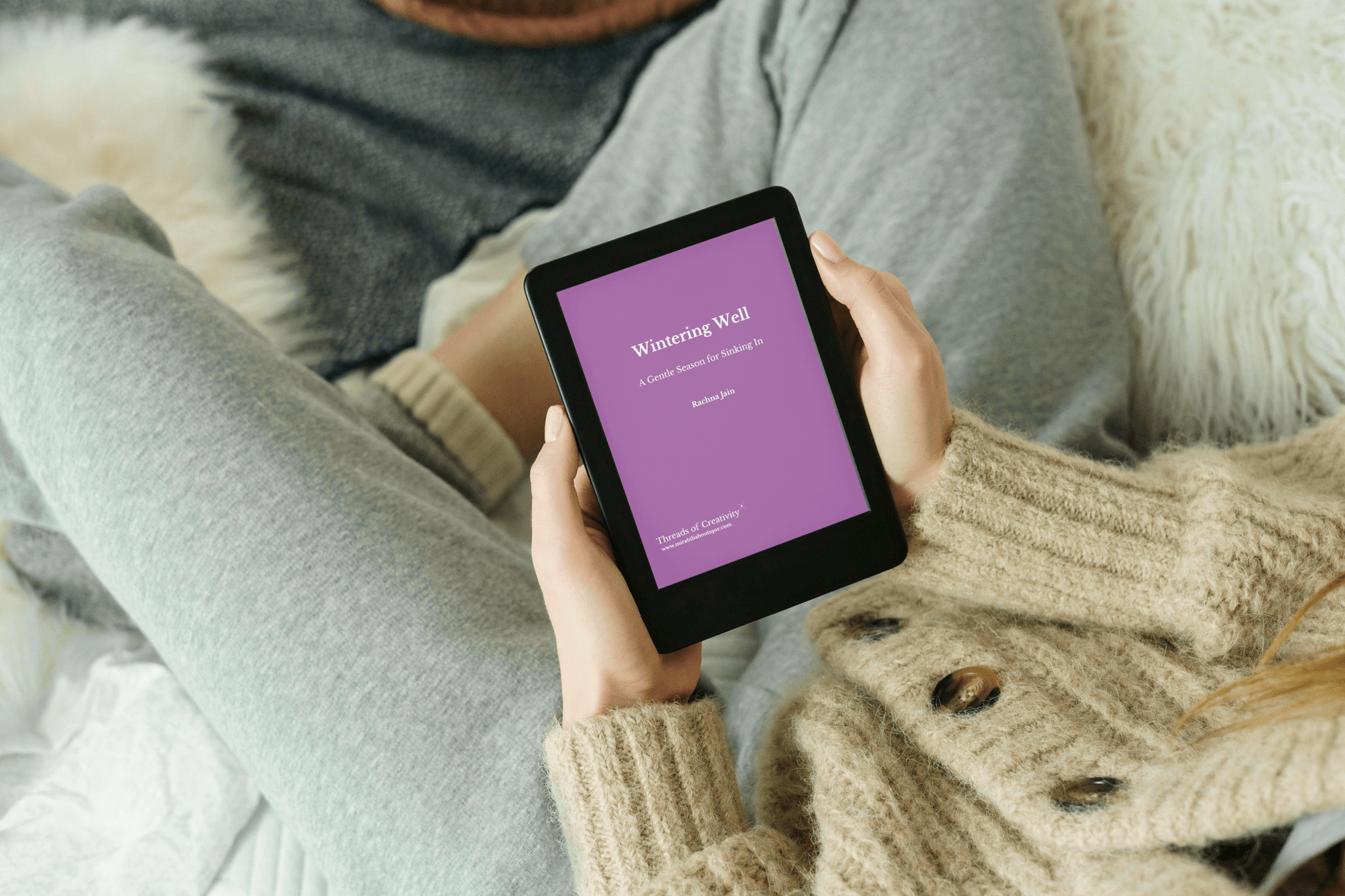 A person in a beige sweater and gray sweatpants sits cross-legged on a couch, holding Mirabilia Boutique’s "Wintering Well Microbook for Quiet, Reflective Seasons"—a perfect companion for winter self-care and inner restoration.