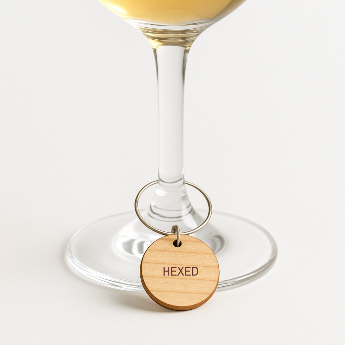 A Witching Hour Wine Charm by Mirabilia Boutique, engraved with “HEXED,” dangles from the stem of a clear wine glass brimming with white wine against a white backdrop—a spellbinding piece of witch decor ideal for your next Halloween gathering.