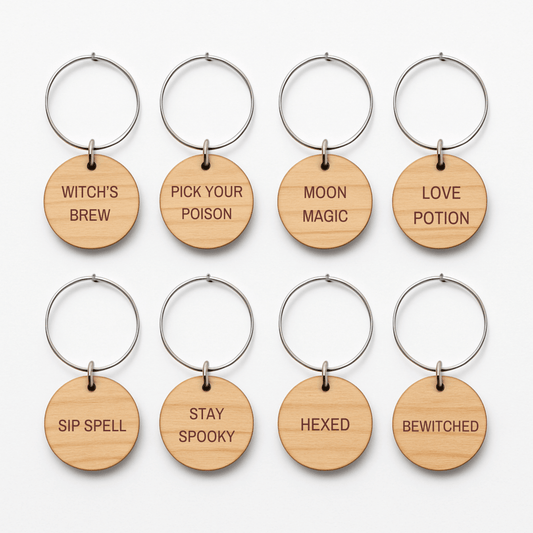 Set of eight Witching Hour Wine Charms by Mirabilia Boutique—round wooden charms with metal rings, each engraved with a distinct Halloween phrase: WITCH’S BREW, PICK YOUR POISON, MOON MAGIC, LOVE POTION, SIP SPELL, STAY SPOOKY, HEXED, and BEWITCHED. Ideal as festive keychains or to enchant your guests as unique wine charms at your next Halloween gathering or for whimsical witchy décor.