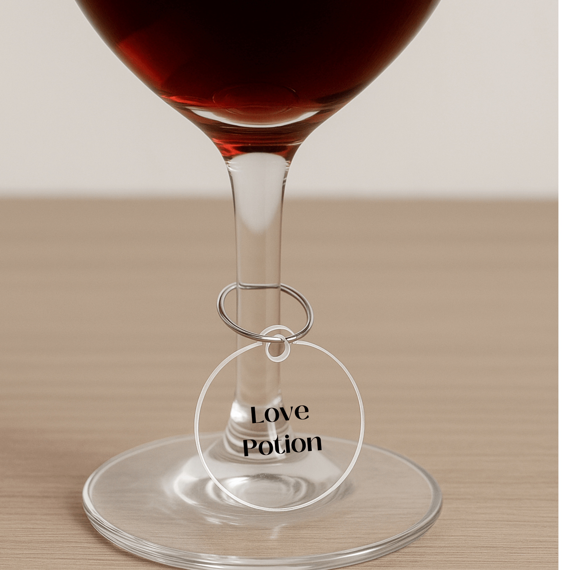A wine glass on wood is adorned with a clear round Witchy Wine Charm from Mirabilia Boutique’s Witchy Wine Charms Set, featuring “Love Potion” in black—an enchanting touch for your Halloween celebration.