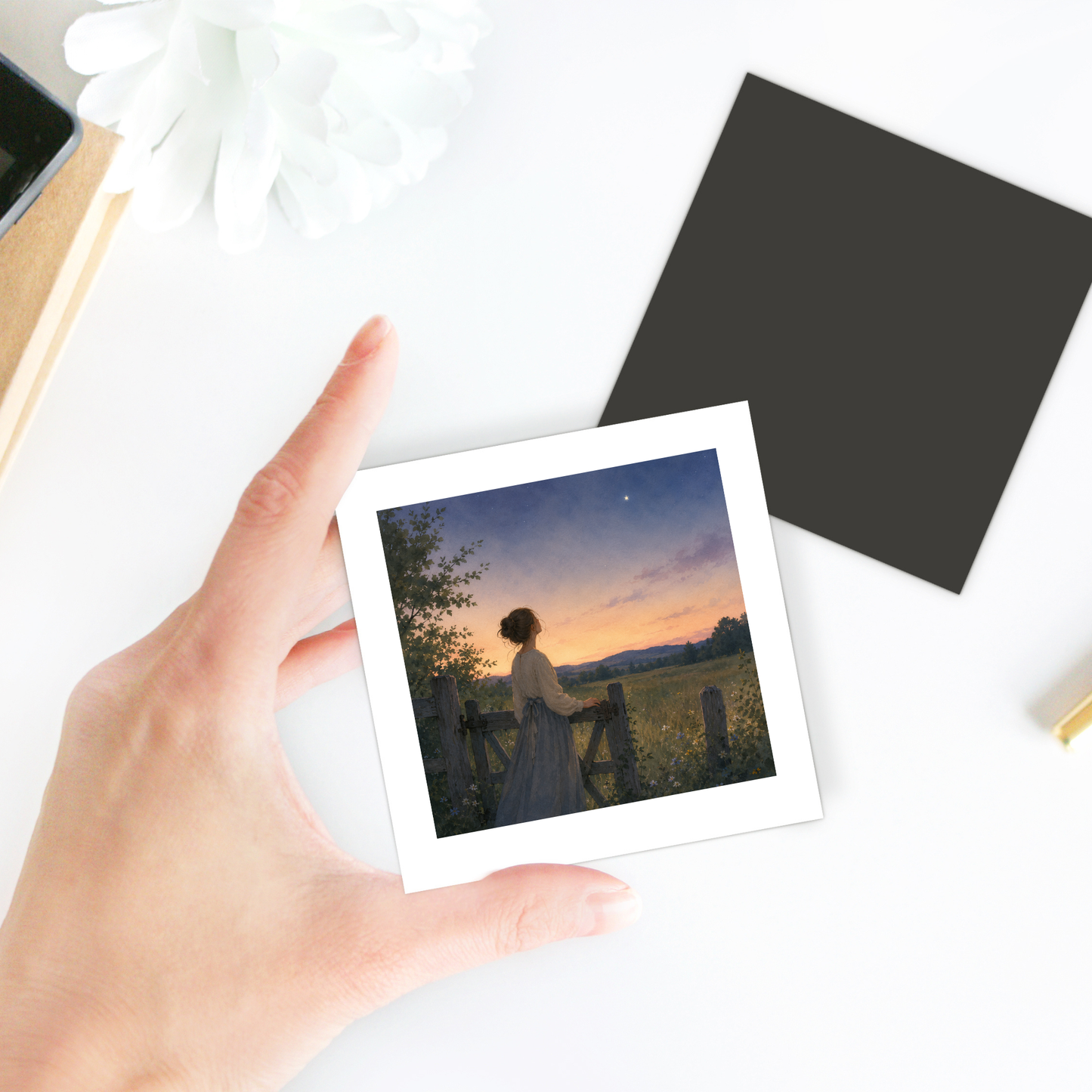 A hand holds the Mirabilia Boutique “Wonder, Always Twilight Landscape Magnet | Nature Art,” featuring a woman in a wildflower meadow at sunset, gazing at the sky. On the table are a white flower, a black square, and a notebook.
