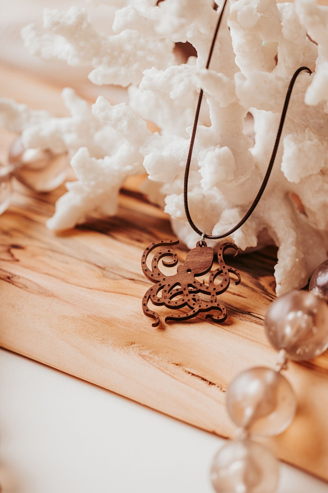 The Wooden Octopus Necklace by Mirabilia Boutique—a nautical pendant for ocean lovers—is shown close-up on a light wooden surface, with white coral and translucent beads providing an elegant, ocean-inspired backdrop.