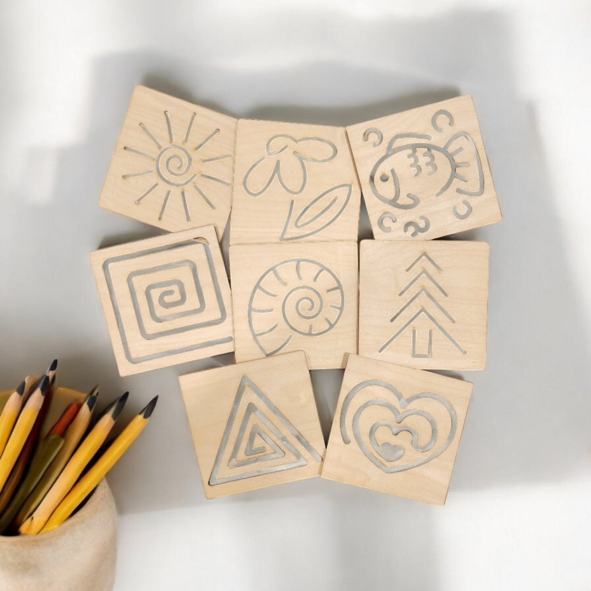 The Montessori Style Tracing Boards Set of 8 by Mirabilia Boutique features wooden boards with etched designs, arranged on a light surface beside colored pencils to help children build fine motor control and pre-writing skills.