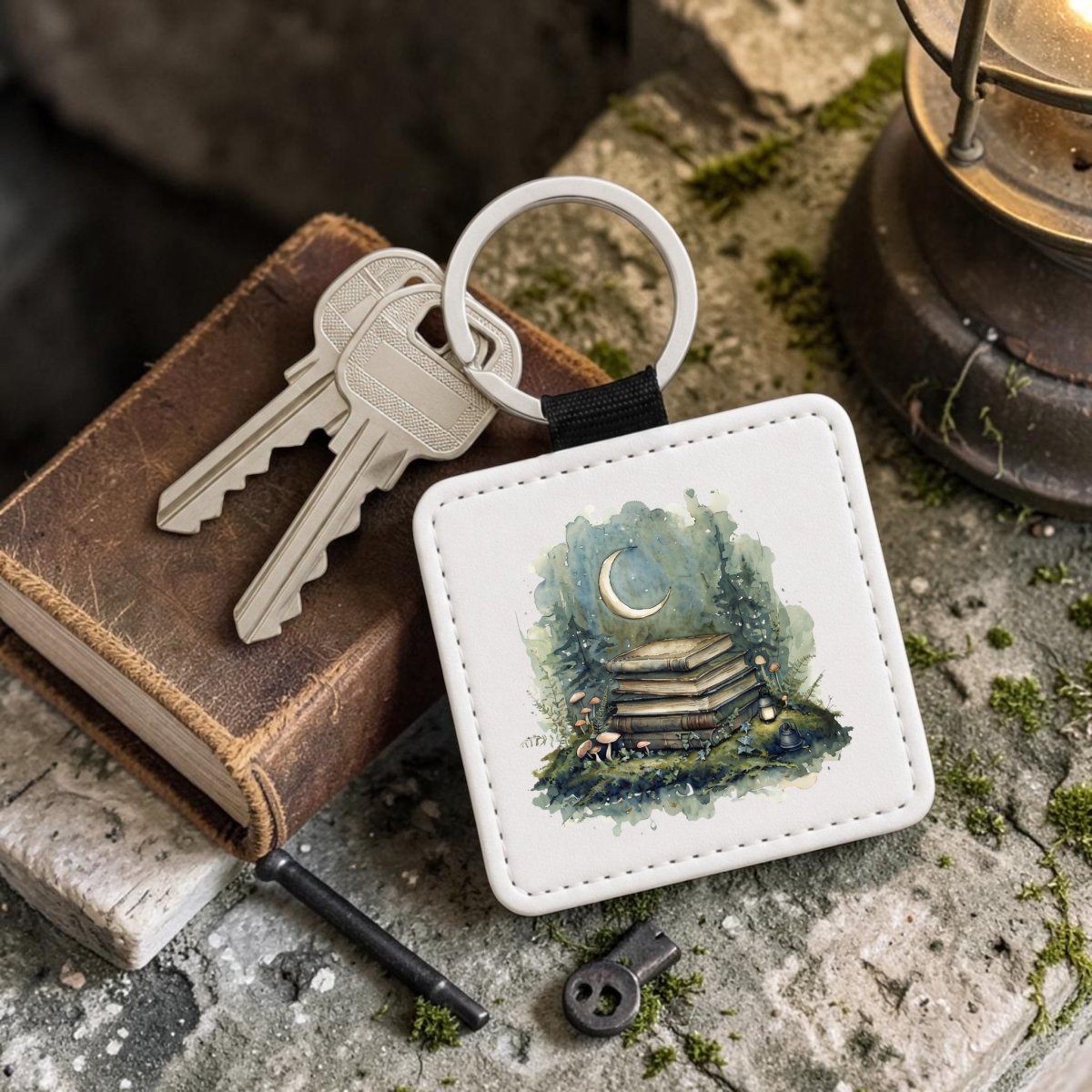 The Bookish Woodland Keychain by Mirabilia Boutique features a whimsical moon and books illustration, accompanied by two silver keys, an old book, a small black key, and a lantern—all resting on a moss-strewn stone surface. This charming cottagecore accessory infuses your everyday essentials with woodland magic.
