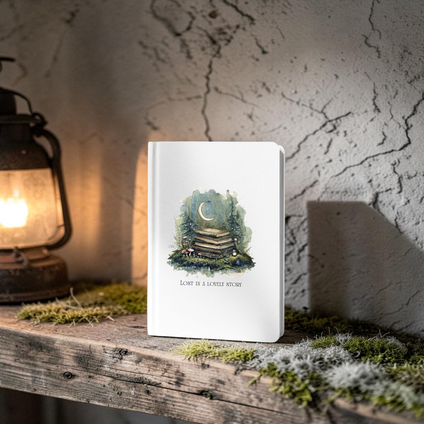 The Lost in a Lovely Story Journal by Mirabilia Boutique—a white hardcover featuring a moon and books illustration along with the words “LOVE IS A LONELY STORY”—stands on a rustic wooden shelf beside a glowing lantern, surrounded by moss and set against a textured stone wall, bringing woodland charm to any space.