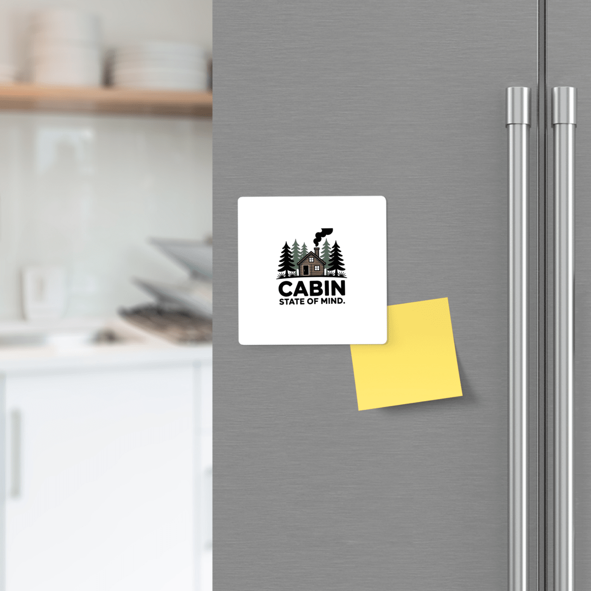 A Mirabilia Boutique "Cabin State of Mind" magnet with a rustic cabin and trees design decorates the fridge door, accented by woodland-themed decor and a yellow sticky note, while kitchen shelves softly blur in the background.