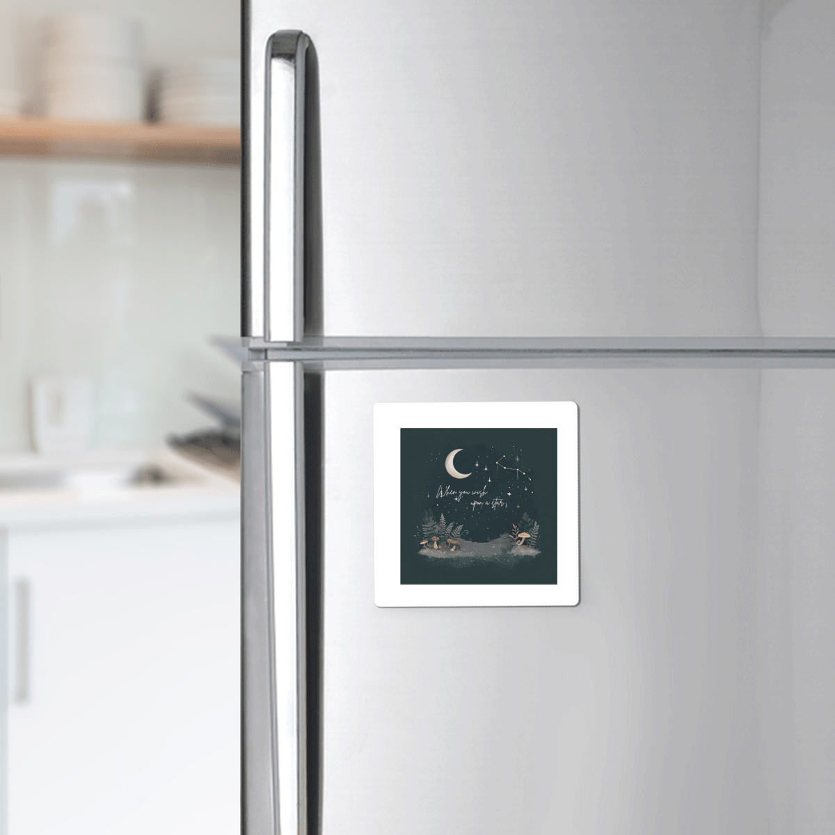 A When You Wish Upon a Star Woodland Magnet by Mirabilia Boutique is displayed on a silver refrigerator, featuring an illustration of two people in boats on nighttime water beneath a crescent moon and stars, with handwritten text. This celestial decor piece stands out against the softly lit, blurred background of the kitchen.