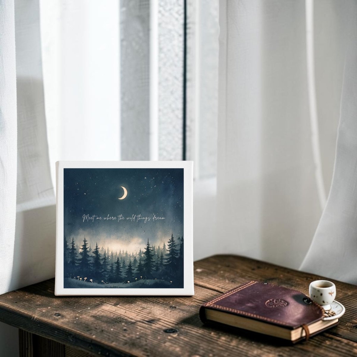 The Meet Me Where the Wild Things Dream Canvas by Mirabilia Boutique rests on a wooden table beside a closed leather journal and a small cup, with sheer white curtains and a window in the background—an enchanting addition to any celestial home decor.