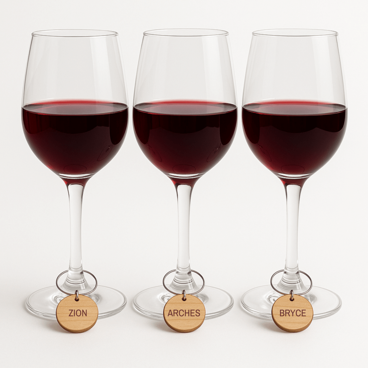 Three wine glasses with red wine are adorned with Mirabilia Boutique’s Southwest National Park Wine Charms Set, labeled "Zion," "Arches," and "Bryce"—perfect national park gifts and a charming touch to any table.