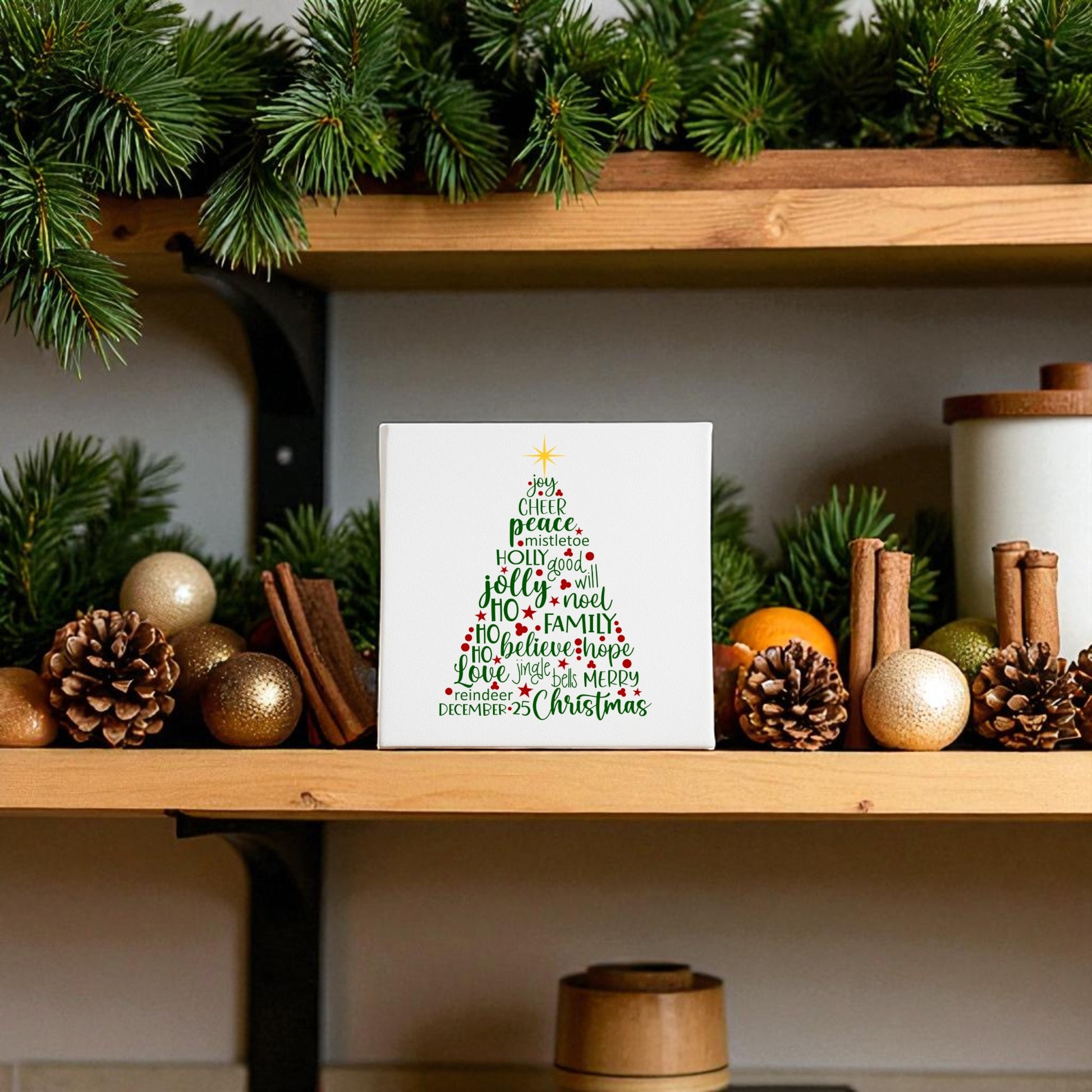 A wooden shelf displays evergreen branches, gold ornaments, cinnamon sticks, and a Mirabilia Boutique Word Art Christmas Tree Canvas 8x8—a festive wall art piece shaped by holiday words for charming Christmas decor.