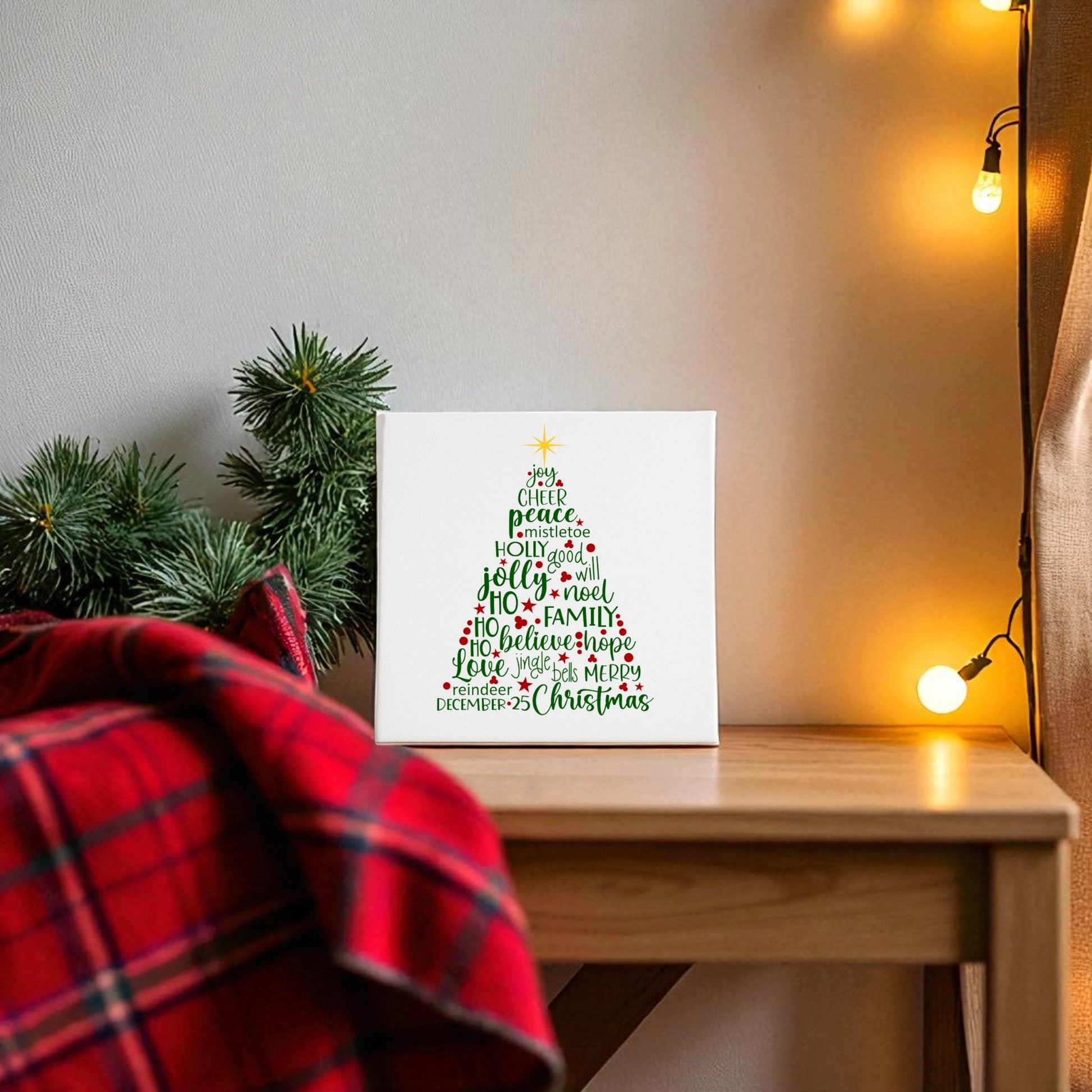 A cozy holiday scene features a wooden table, red plaid blanket, pine branches, and string lights, highlighted by Mirabilia Boutique's Word Art Christmas Tree Canvas (8x8) with festive words in colorful text.