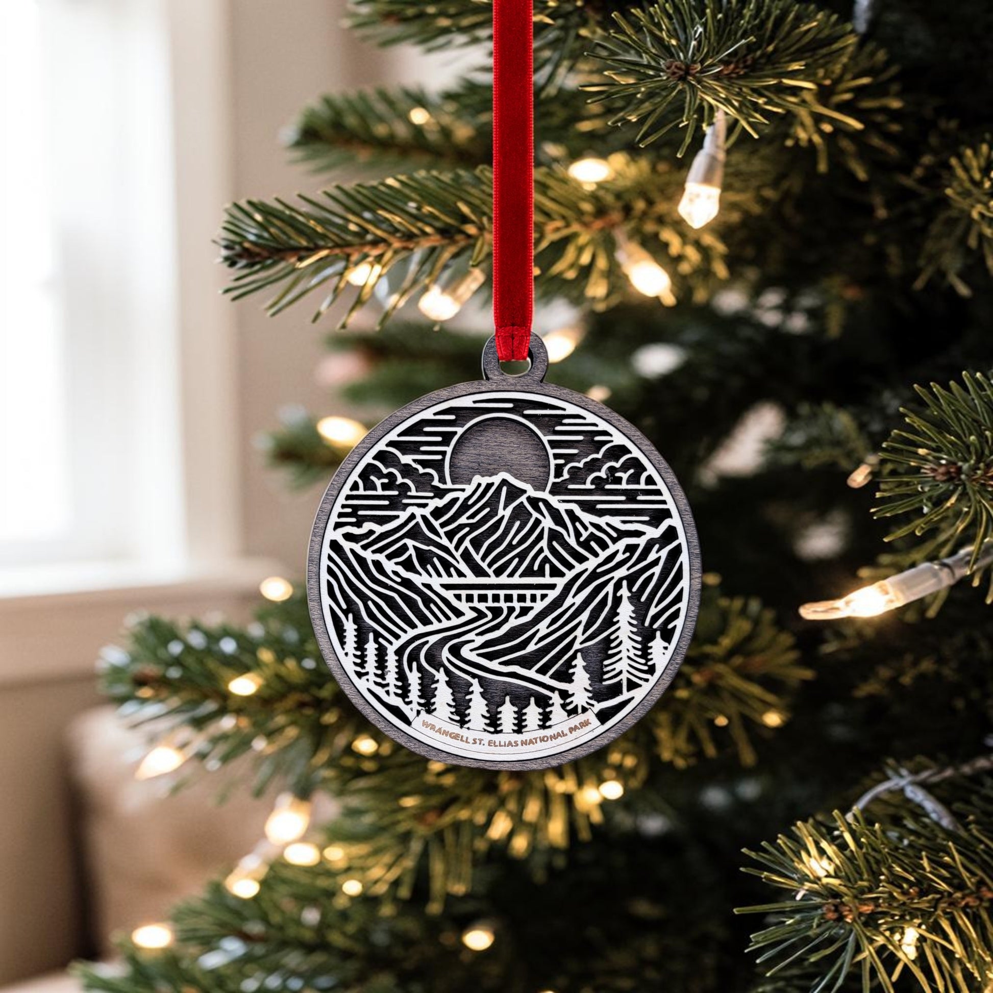 The Wrangell-St. Elias National Park Line Art Christmas Ornament by Mirabilia Boutique—a handcrafted wooden piece showcasing a mountain and river motif inspired by Alaska's wilderness—hangs from a red ribbon on a Christmas tree adorned with white string lights.