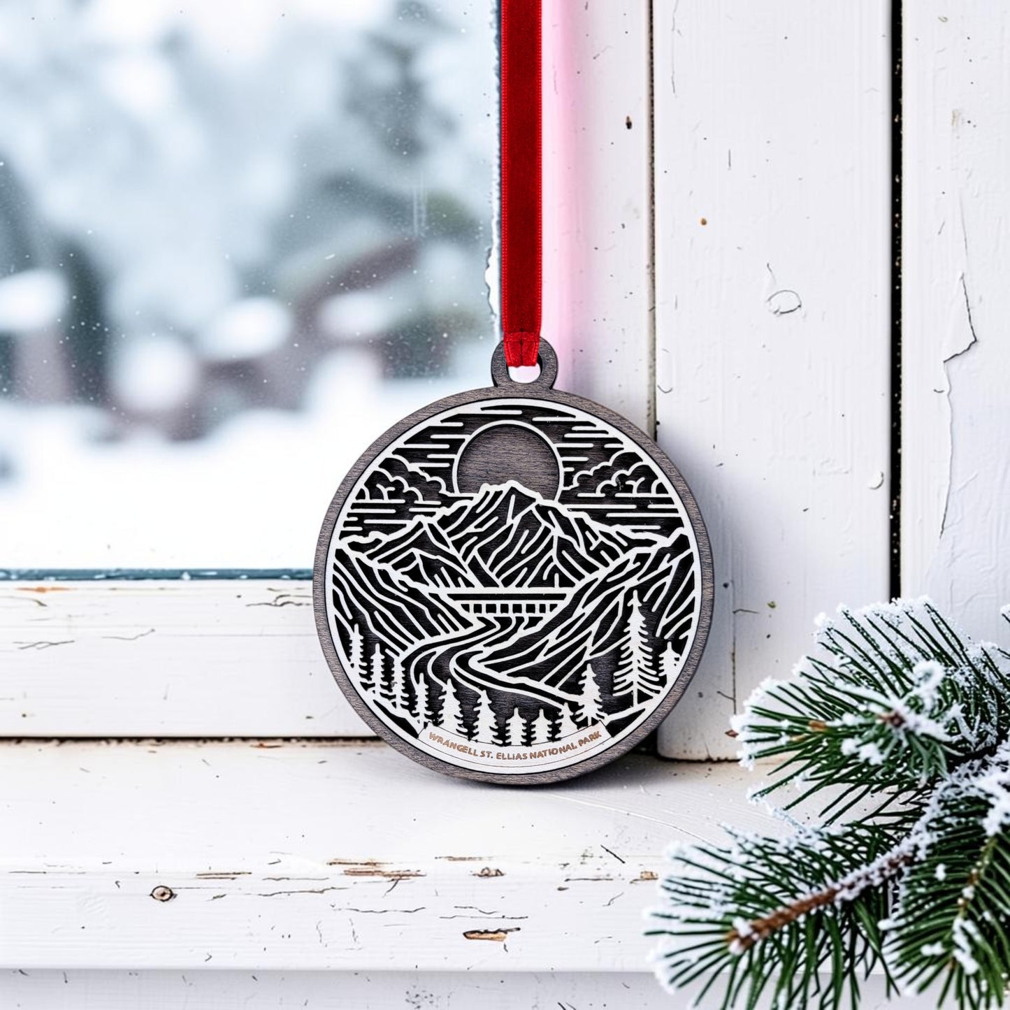 A round, handcrafted Wrangell-St. Elias National Park Line Art Christmas Ornament from Mirabilia Boutique hangs by a red ribbon on a snowy window. Featuring an intricate mountain and forest scene that captures the beauty of the Alaska wilderness, pine branches dusted with snow are visible in the lower right corner.