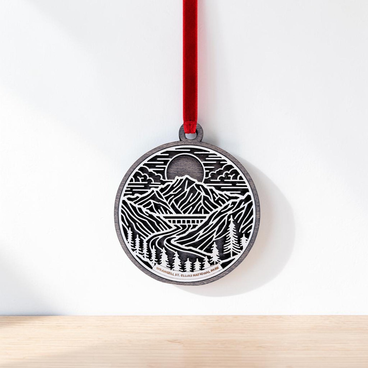 The Wrangell-St. Elias National Park Line Art Christmas Ornament by Mirabilia Boutique is a round, handcrafted wooden ornament featuring a black and white mountain landscape design. Hanging from a red ribbon, it depicts mountains, trees, a winding river, and a sun or moon in the sky—capturing the spirit of Alaska wilderness decor inspired by themes from Wrangell-St. Elias National Park.