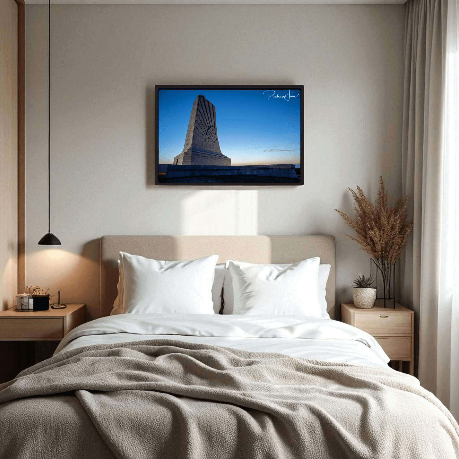 A cozy bedroom features a neatly made bed, two bedside tables with lamps, a vase of dried plants, and the Mirabilia Boutique Wright Brothers Monument Print | First in Flight Wall Art hanging above the bed.