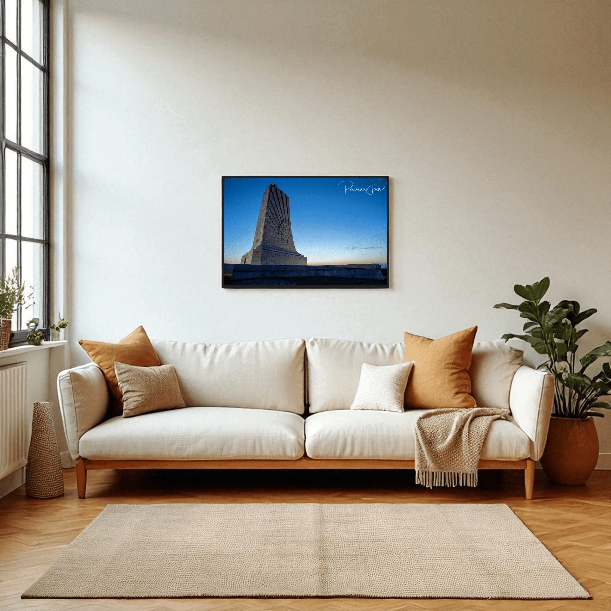 A modern living room with a white sofa, brown and beige pillows, a large indoor plant, and the Mirabilia Boutique "Wright Brothers Monument Print | First in Flight Wall Art" framed above the couch. Sunlight pours in from tall windows on the left.