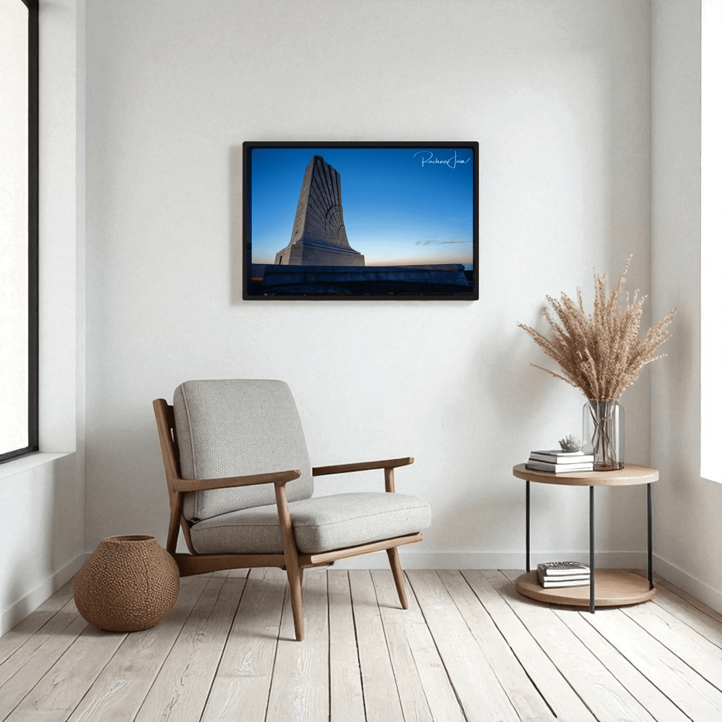 A modern living room with a cushioned chair, woven pouf, and round side table holding a vase of dried grass. On the wall hangs Mirabilia Boutique’s First in Flight Wright Brothers Monument Print. Light wood floors and white walls complete the look.